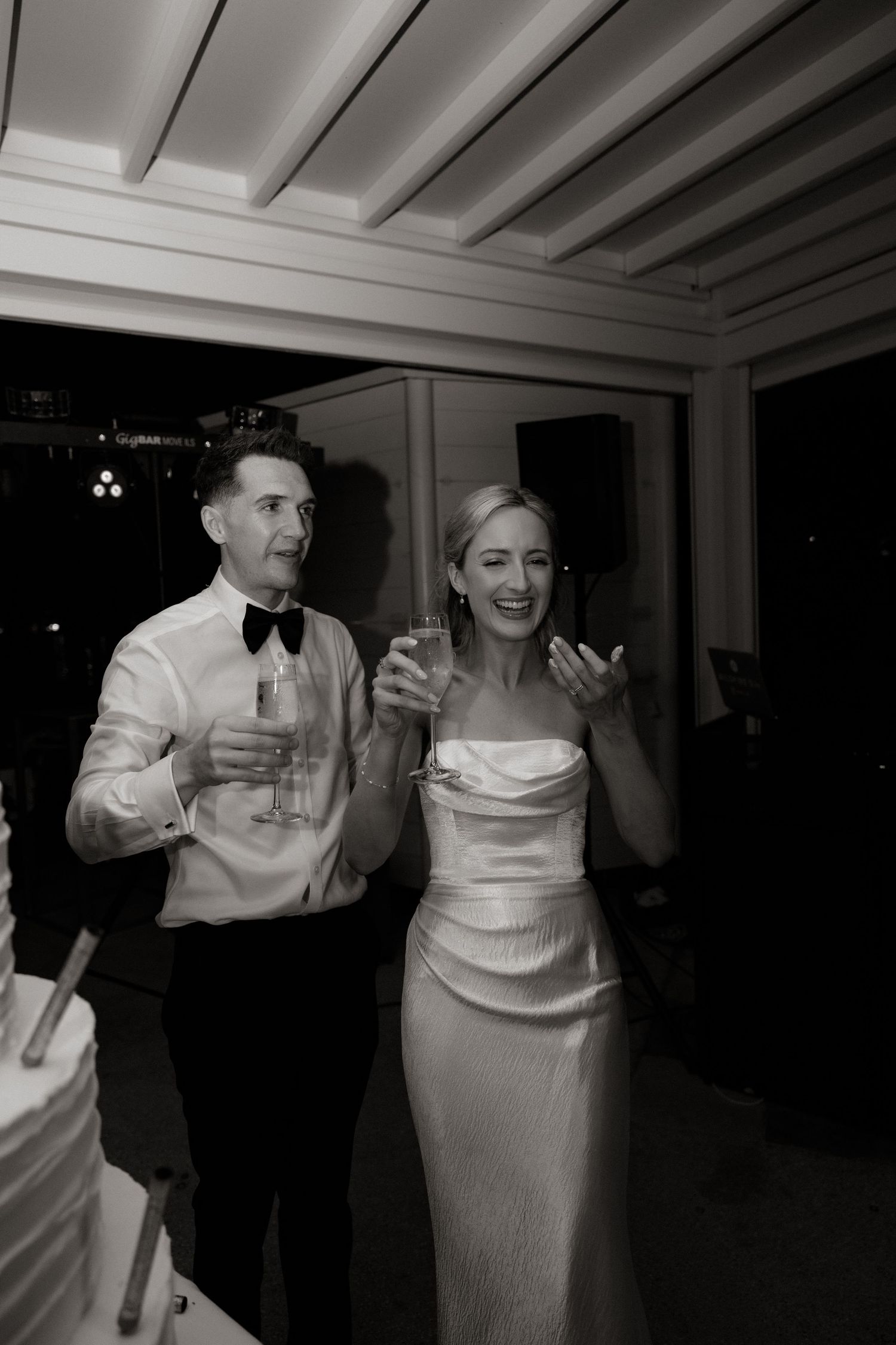 A wedding couple raises champagne glasses in celebration next to their white tiered cake.