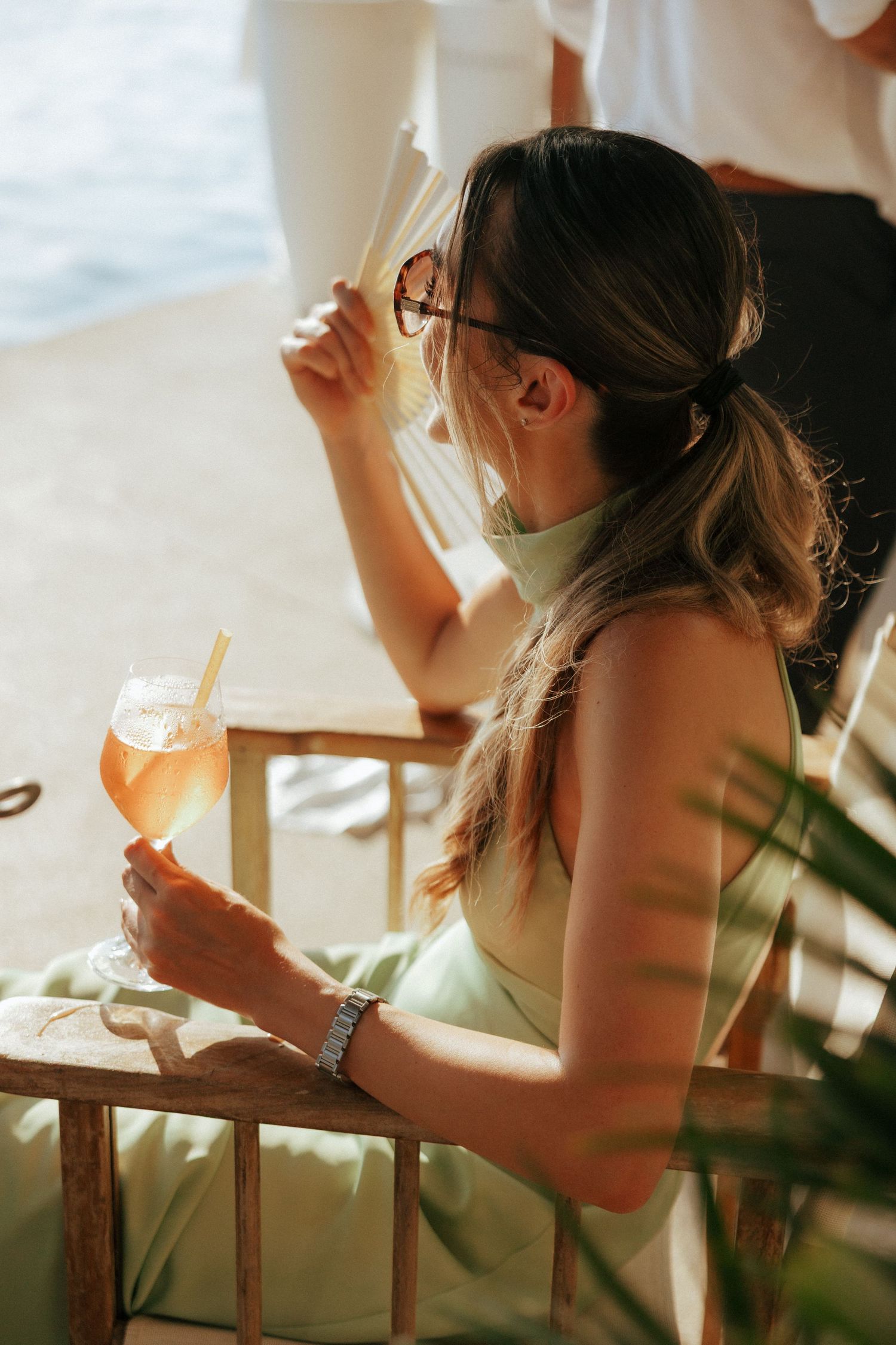 Woman in glasses sips from orange cocktail near window during golden hour while relaxing at home.