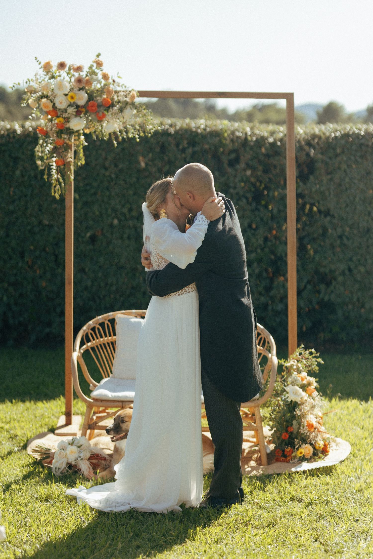 Couple embracing under floral arch with rattan chair and garden backdrop at outdoor wedding.