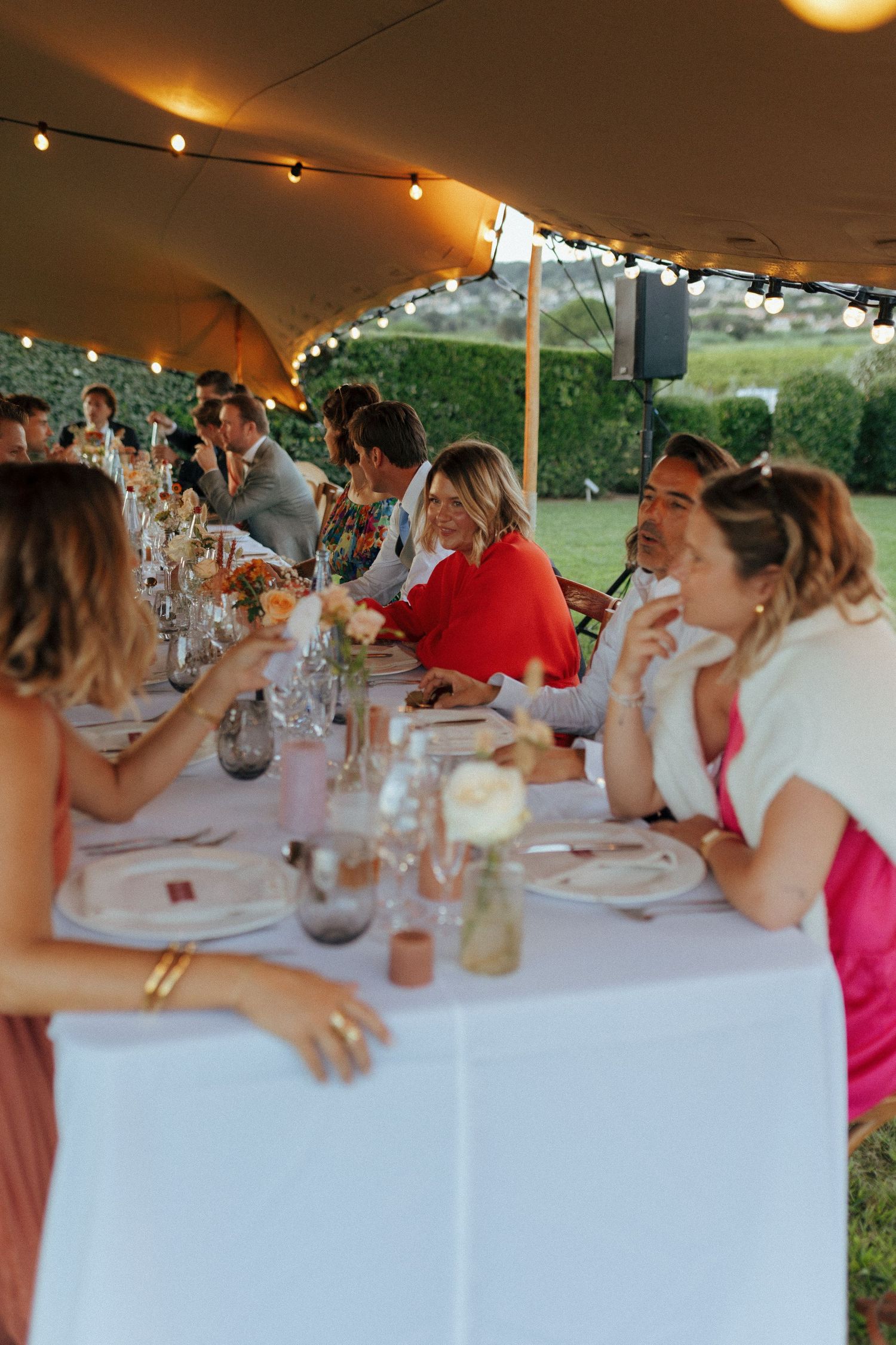 Wedding guests dine under a tent with string lights at long tables set for an outdoor reception.