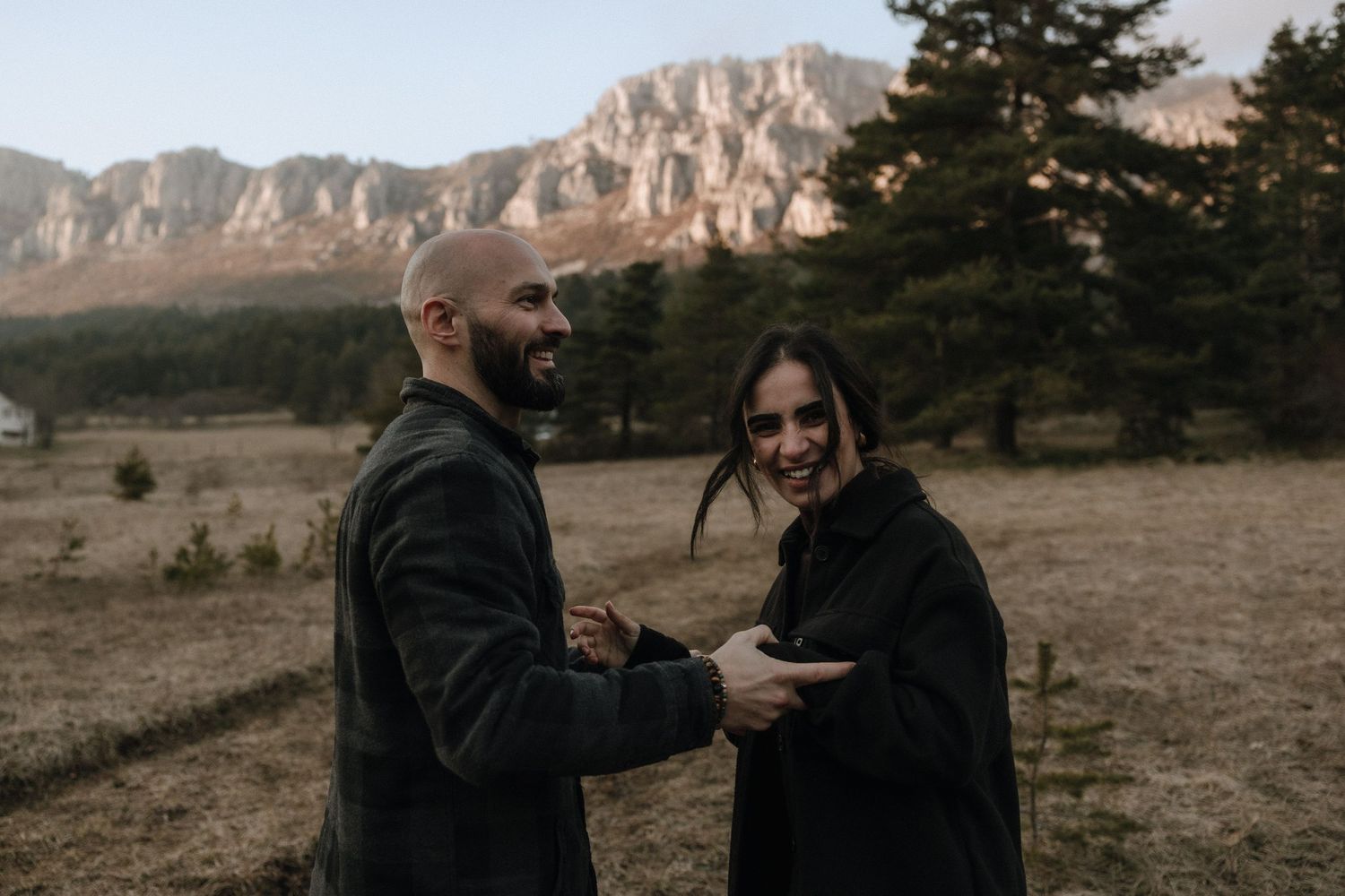 Two people share a laugh in front of majestic mountains and evergreen trees during golden hour.