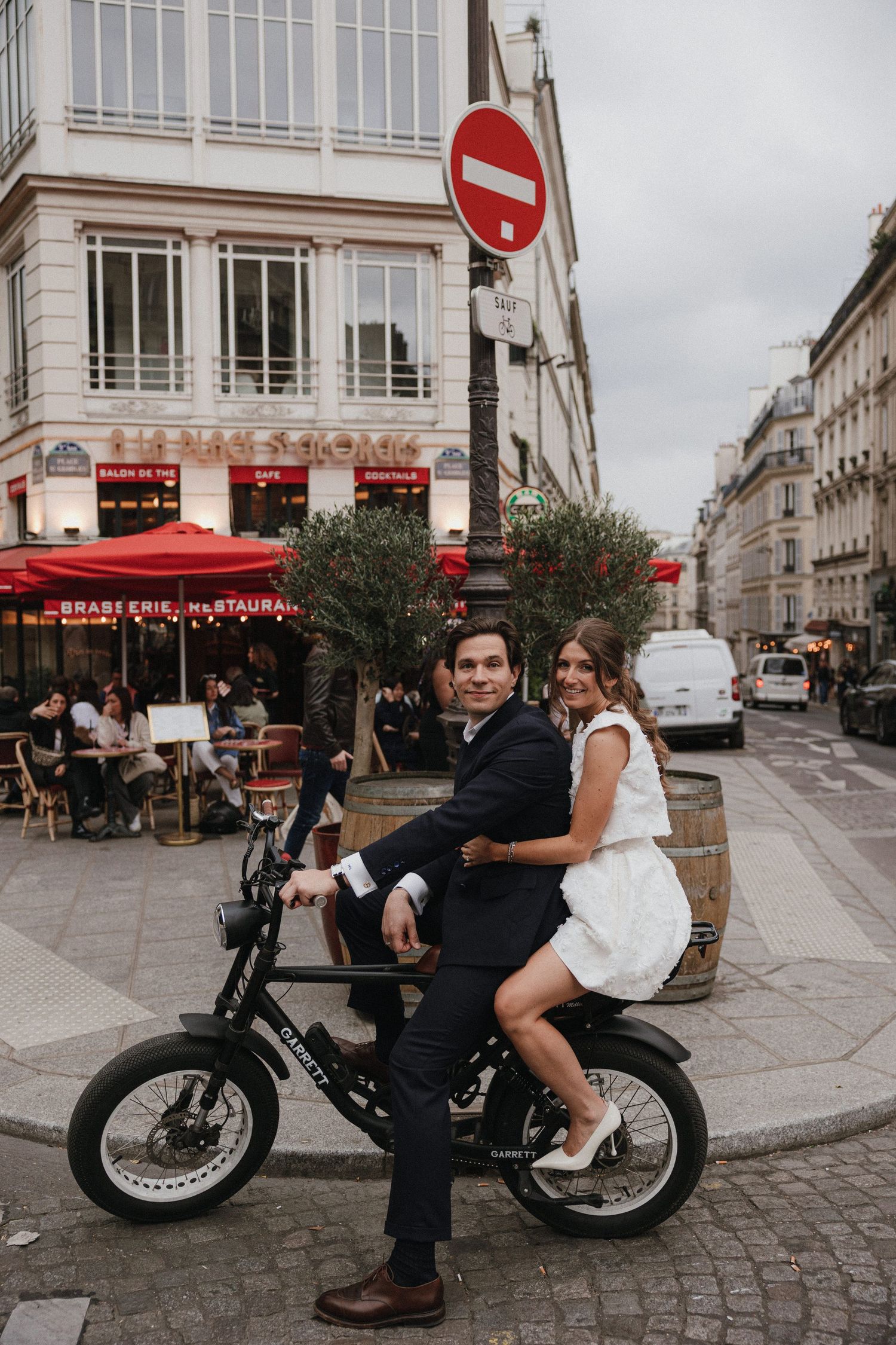 A couple on an electric bike outside a Parisian cafe with red awning on a cobblestone street corner.