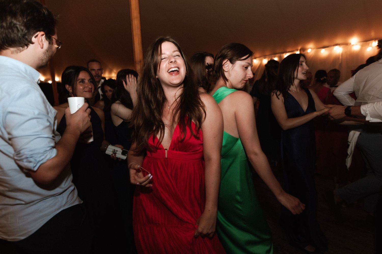 Lively wedding reception dance party with guests in colorful dresses under warm string lights.