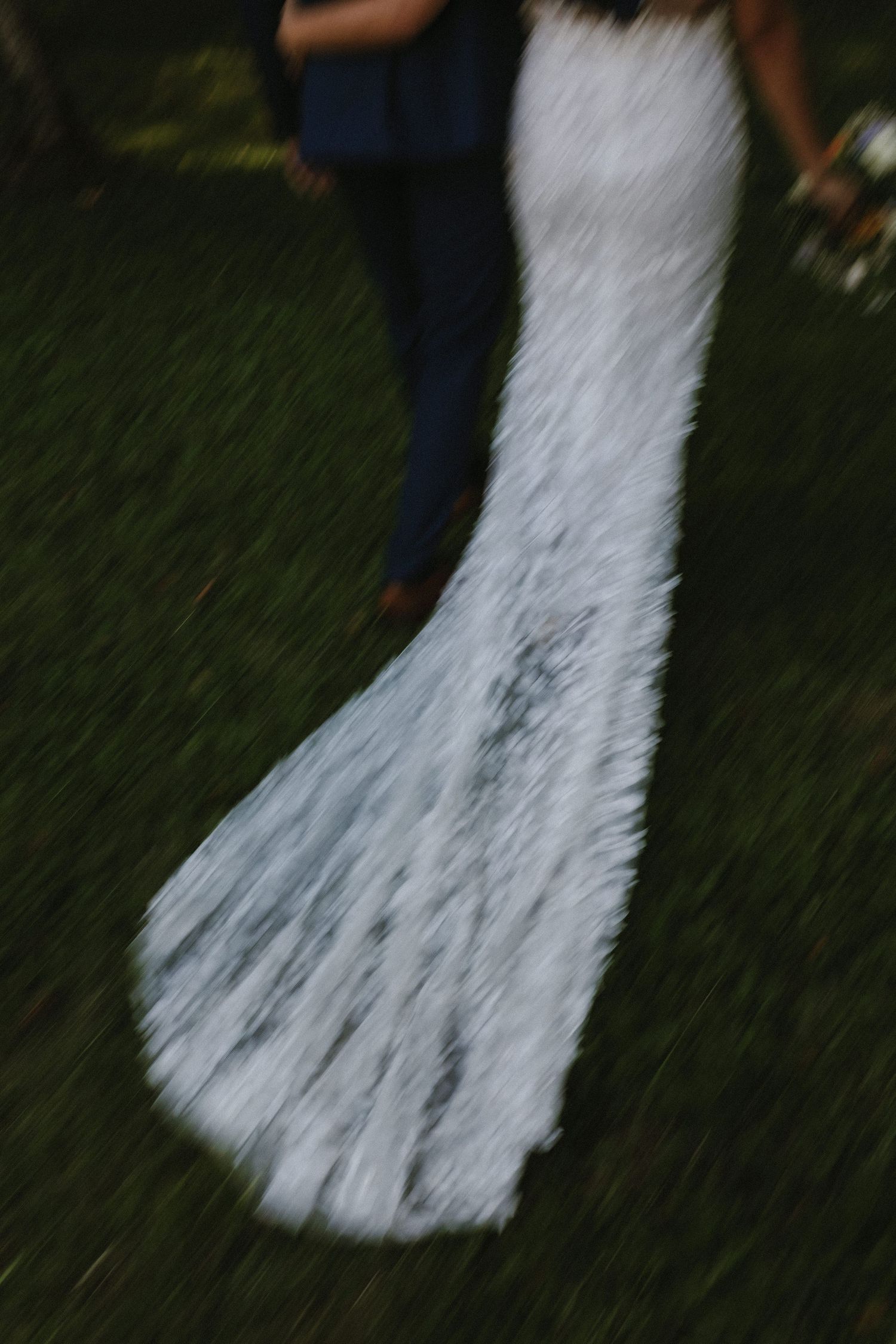A motion-blurred white lace wedding dress train trails on green grass, creating an ethereal effect during an outdoor ceremony.