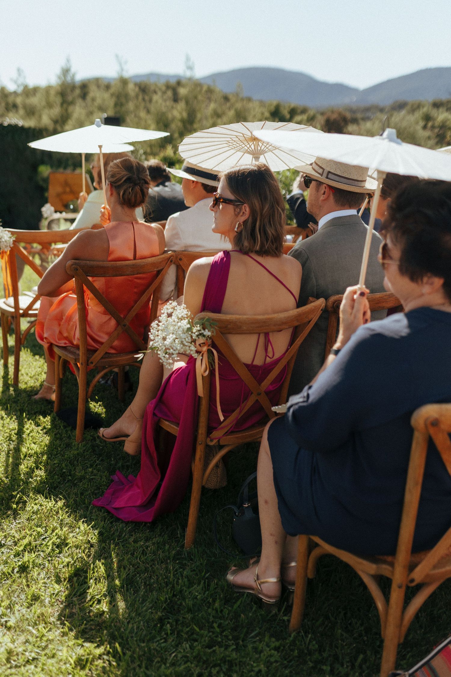 Wedding guests seated in wooden chairs with parasols at an outdoor ceremony with mountain views.