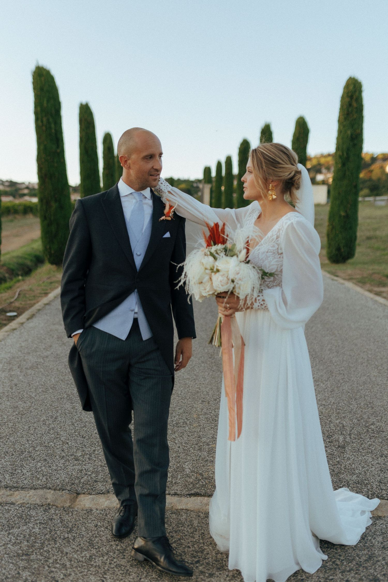 Elegant wedding couple strolls down cypress tree-lined path with bridal bouquet.