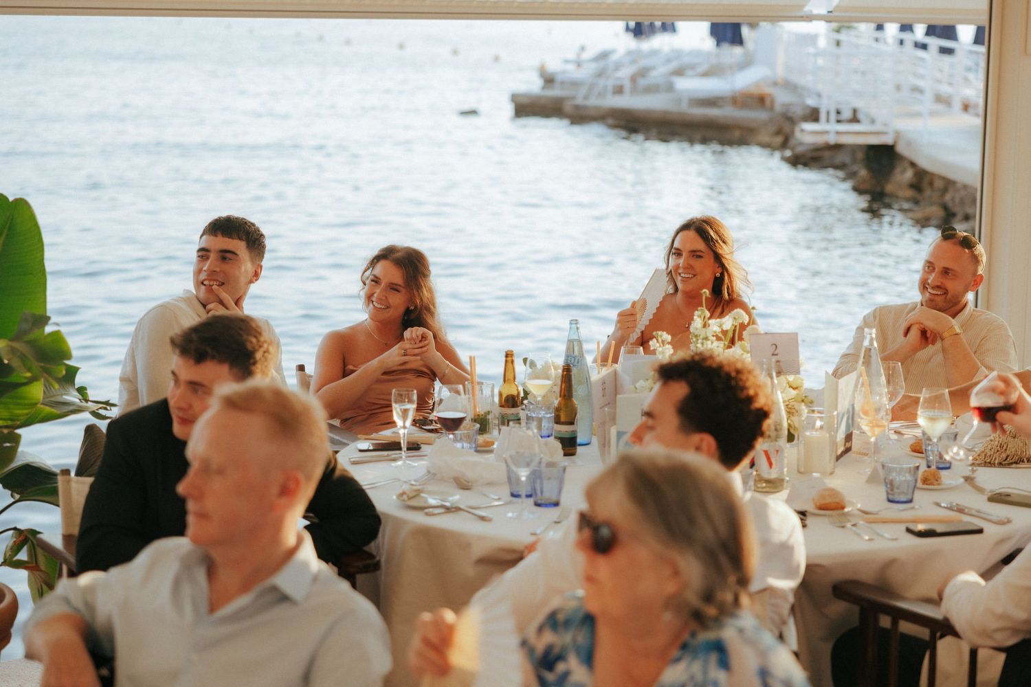Wedding guests enjoy an elegant outdoor dining experience at a waterfront venue with boats in the background.