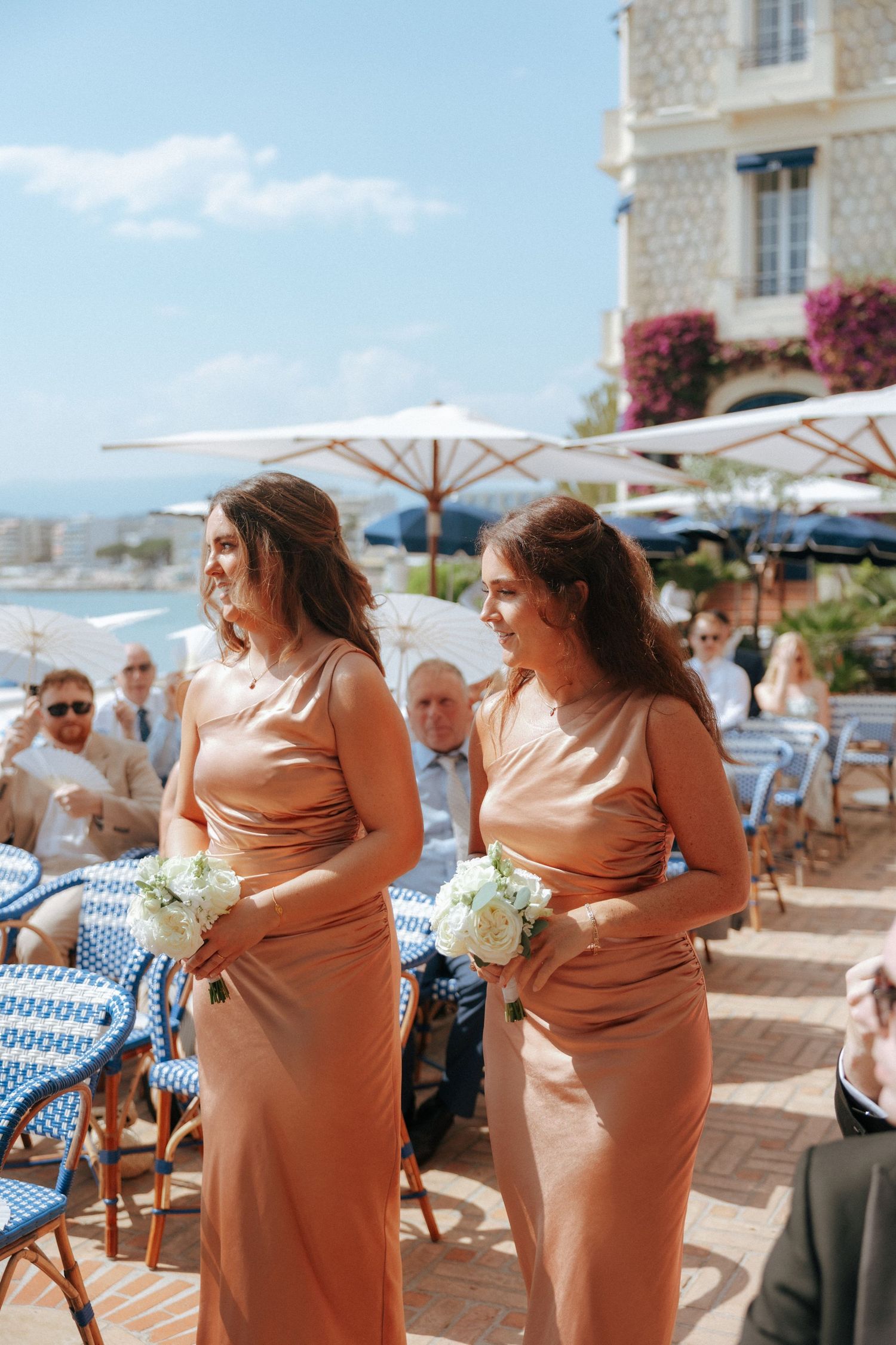 Two bridesmaids in peachy-colored dresses hold white bouquets at an outdoor seaside venue with umbrellas.