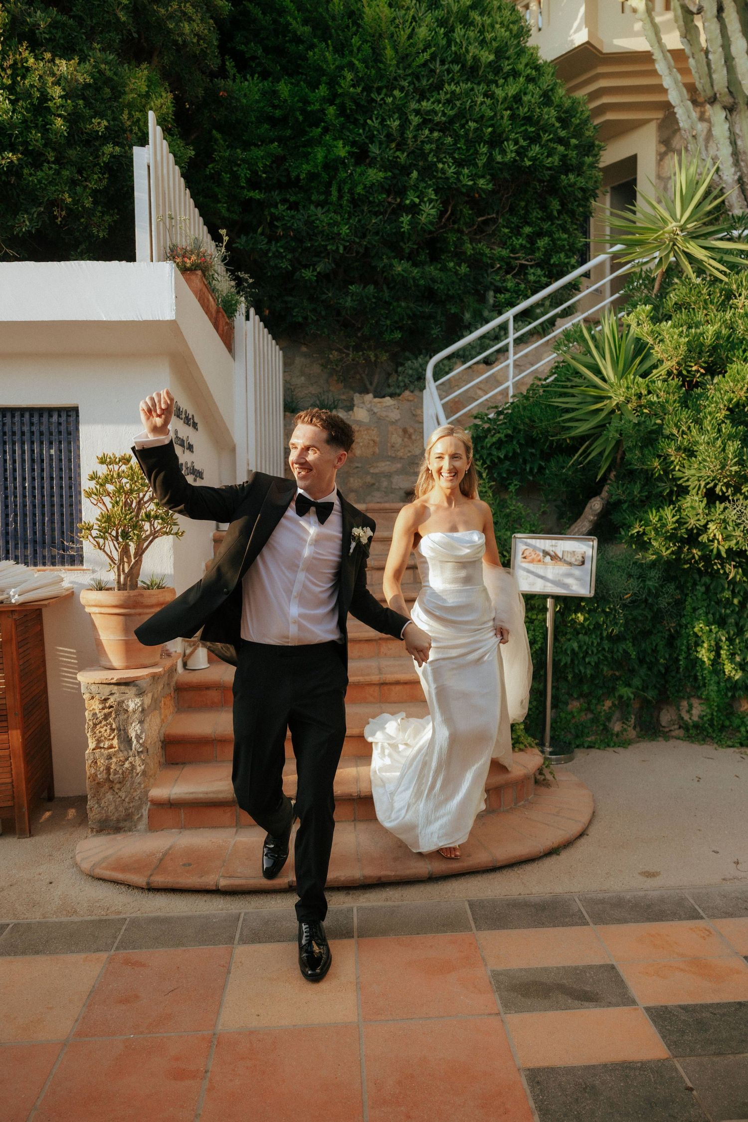 A newlywed couple joyfully exits down outdoor steps at their wedding venue, with lush greenery in the background.