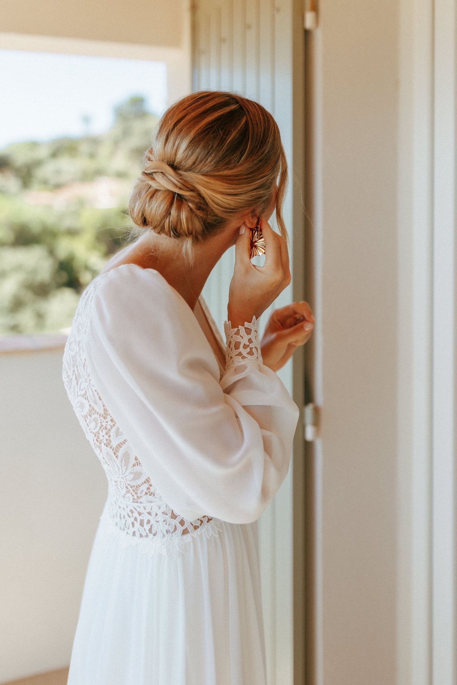Elegant bridal gown with lace details and flowy sleeves shown from behind as bride adjusts earring near window.