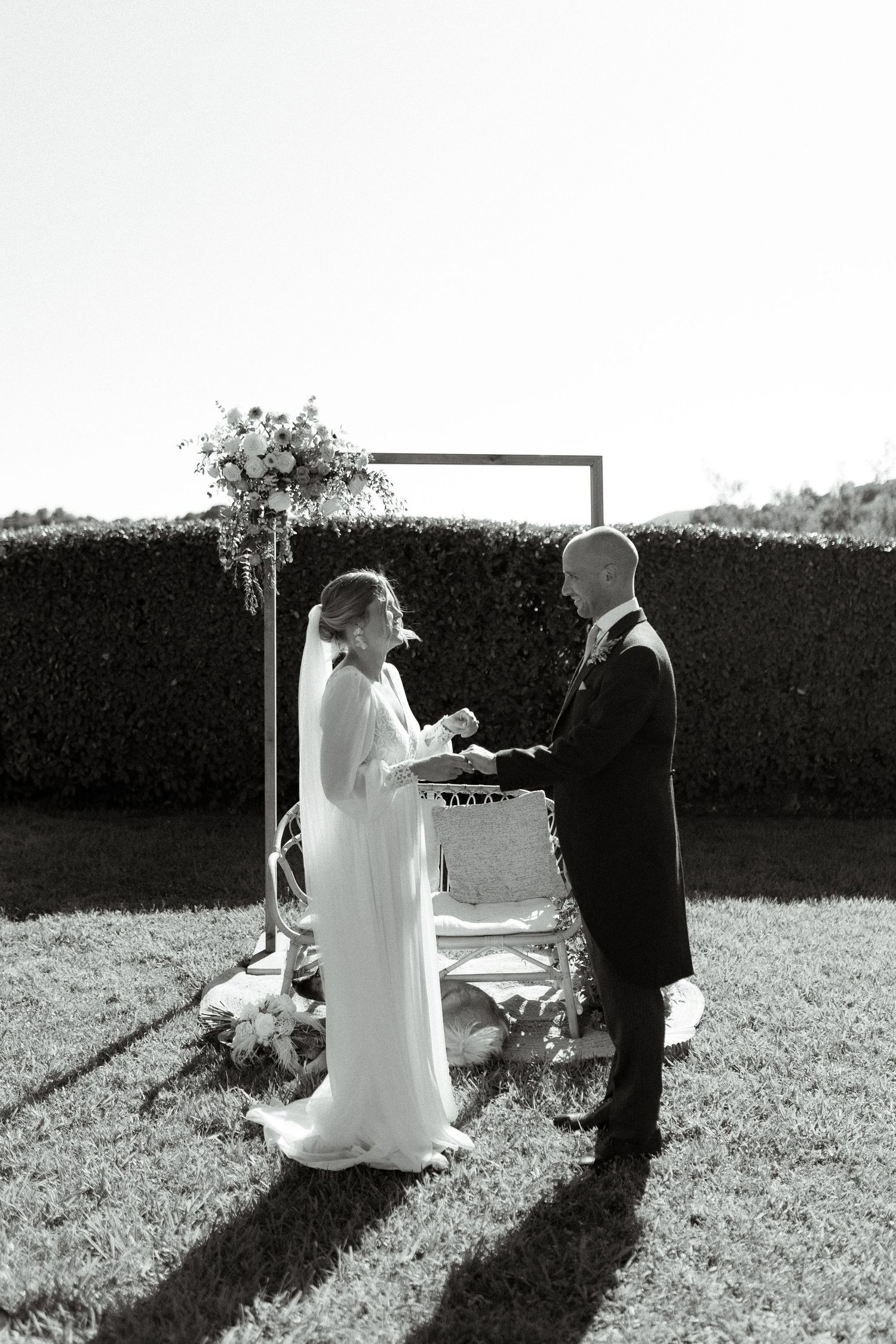 A wedding ceremony taking place in front of a minimalist arch against a hedge backdrop.