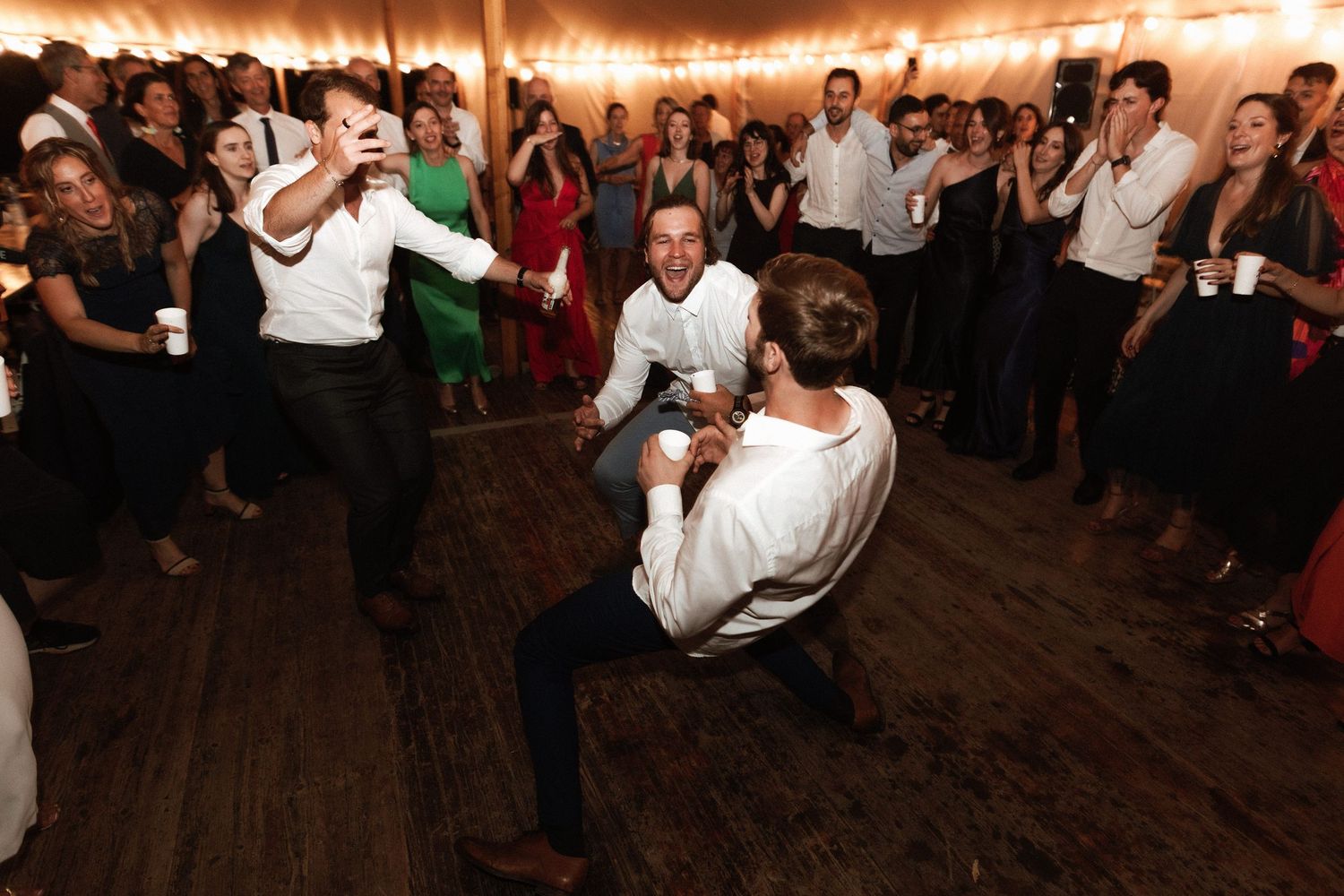 Party guests dance energetically on a wooden dance floor at a dimly lit event space surrounded by onlookers.