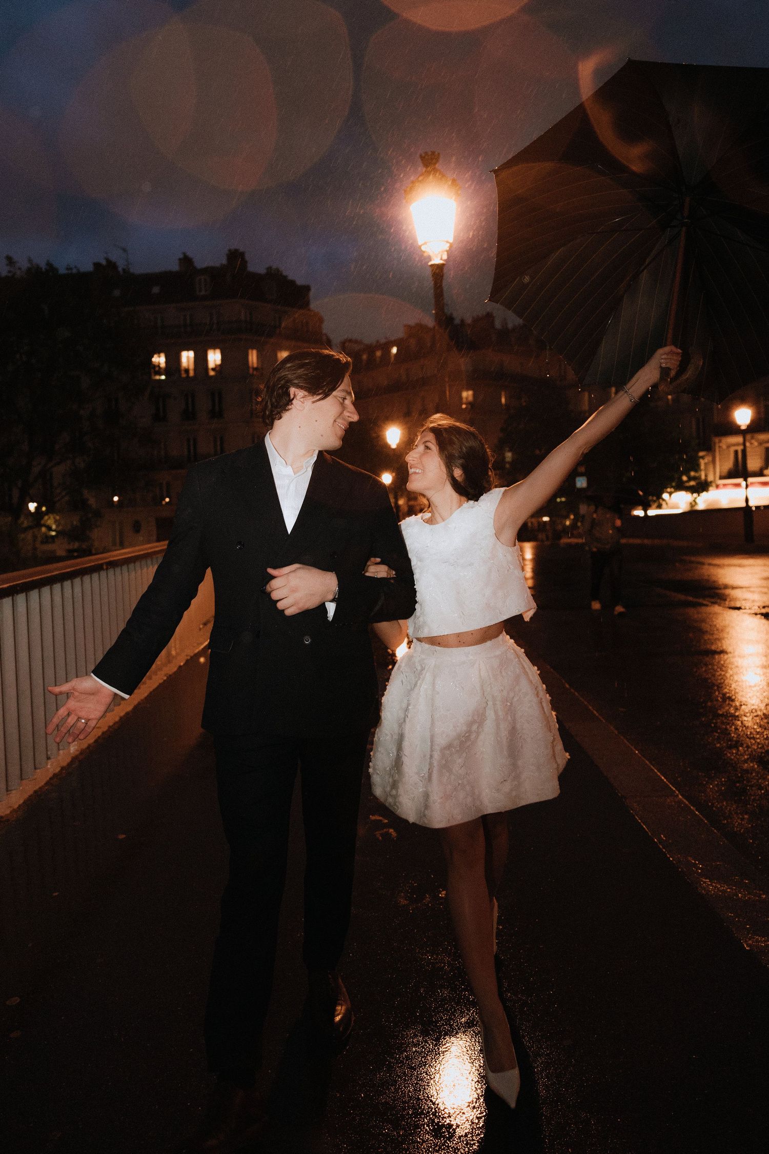 A couple dances joyfully at night on a rainy street in Paris with glowing lampposts illuminating the romantic scene.