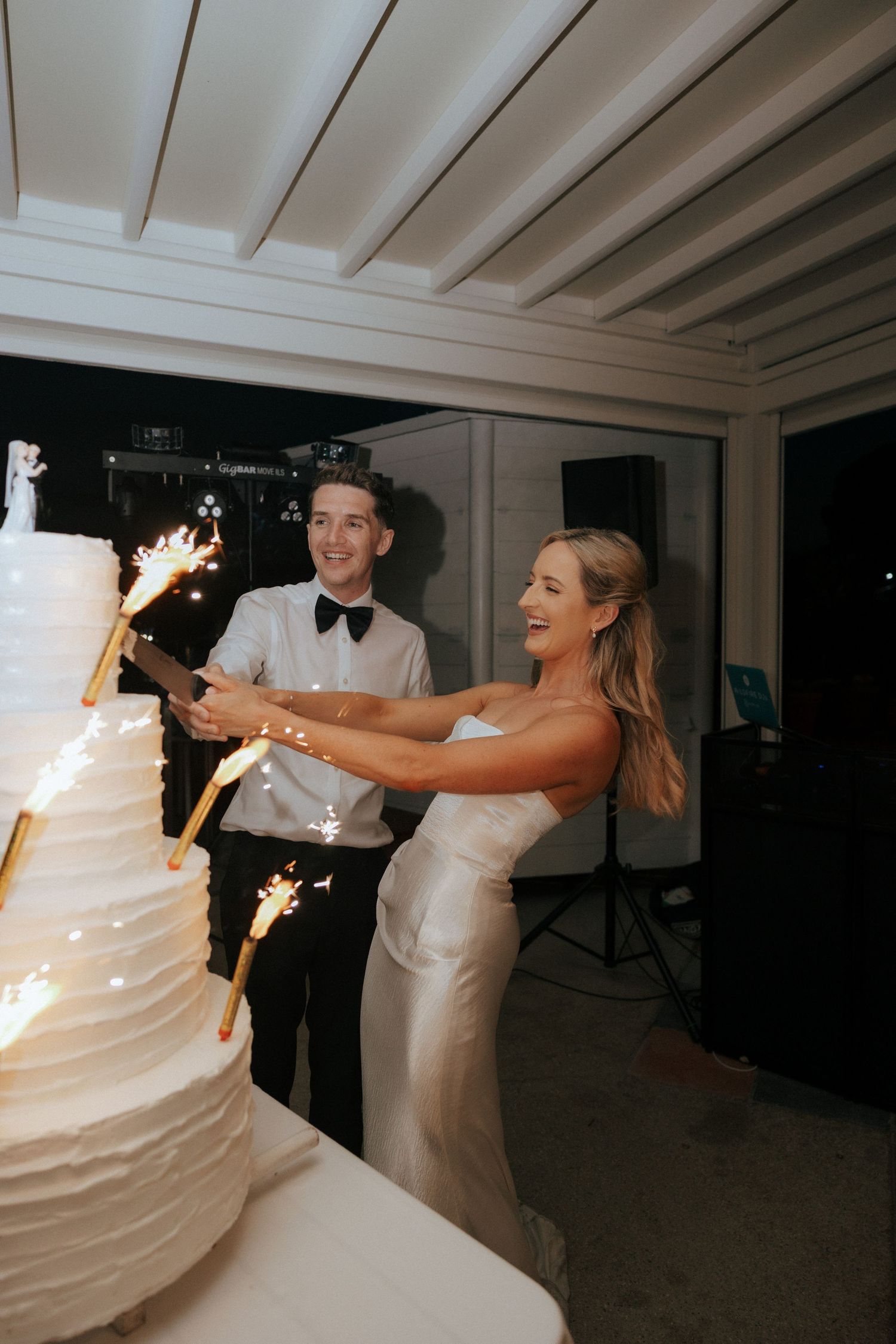 Newlyweds share a joyful moment while cutting their tall white wedding cake adorned with sparklers.