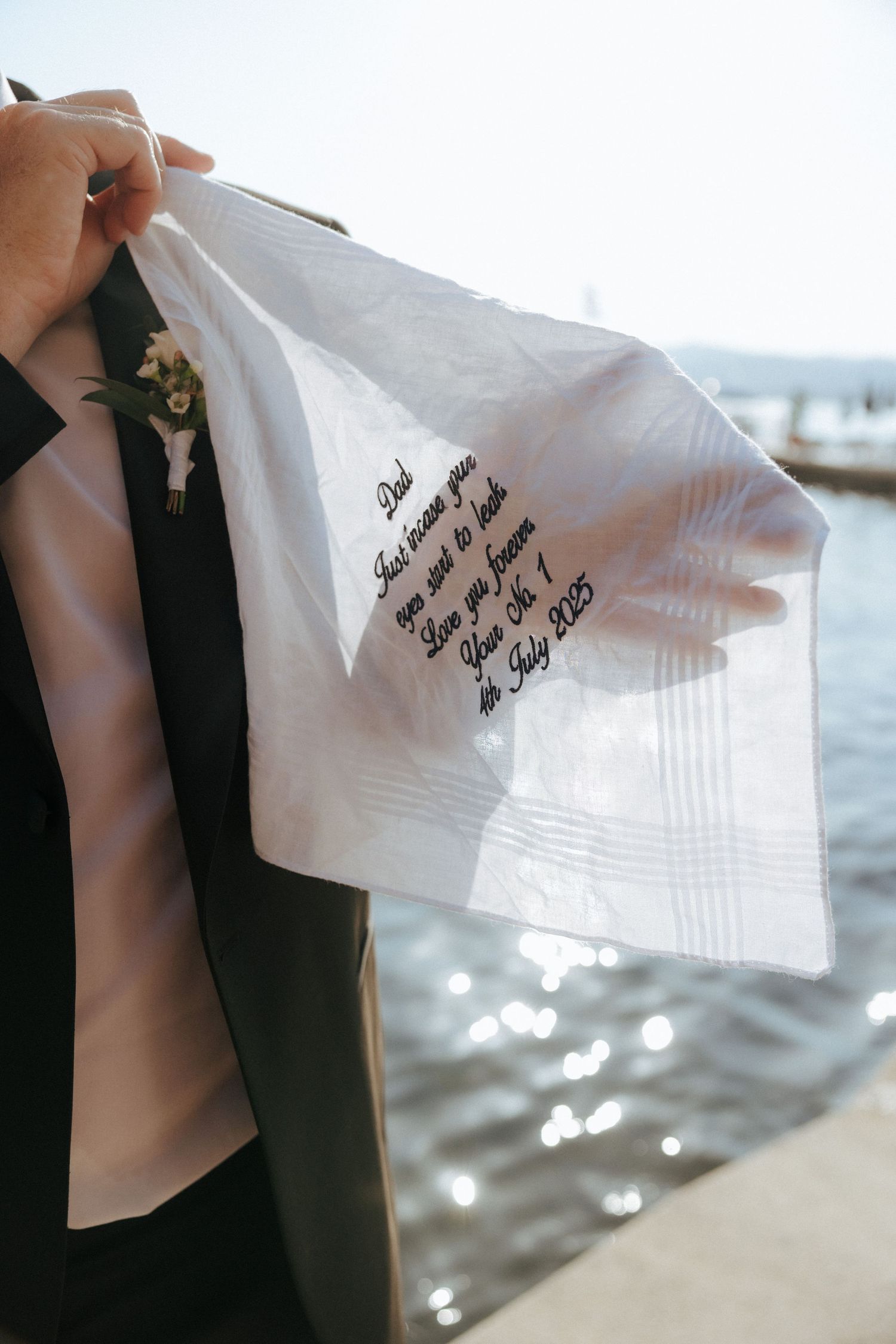 A delicate white handkerchief with handwritten text held against a sparkling waterfront background.