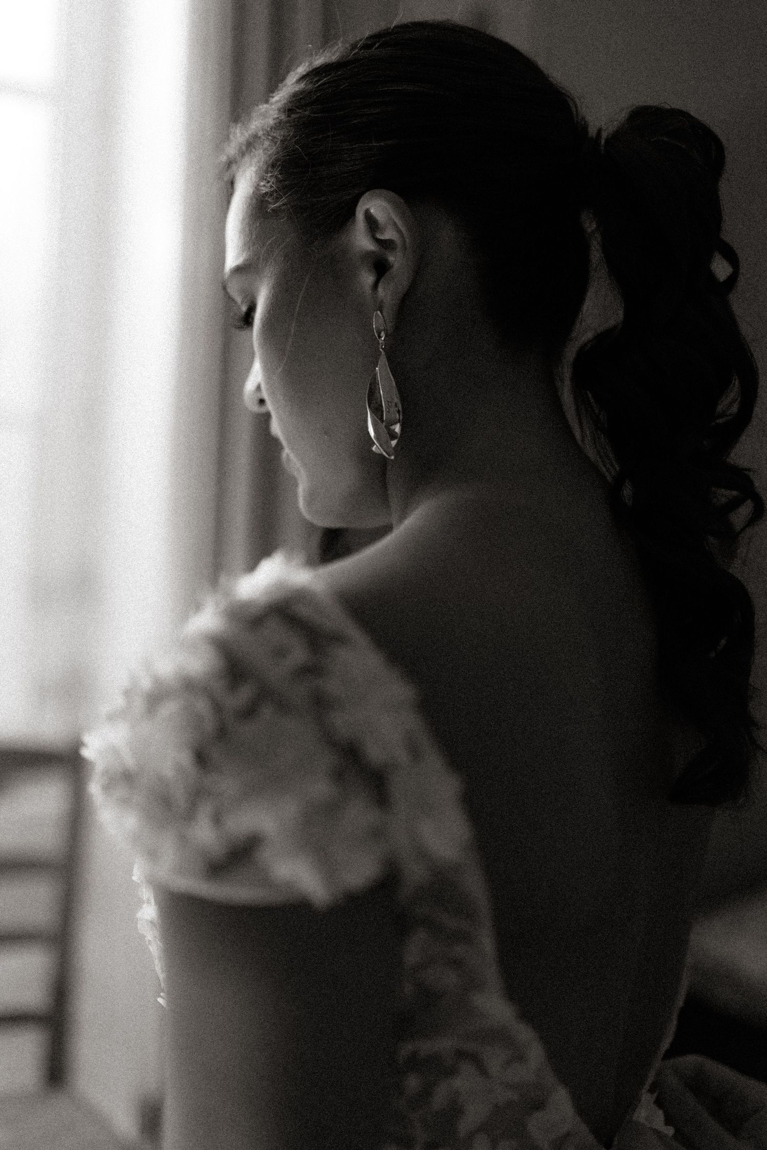 Black and white portrait from behind showing an updo hairstyle, dangling earring, and lace off-shoulder wedding dress.