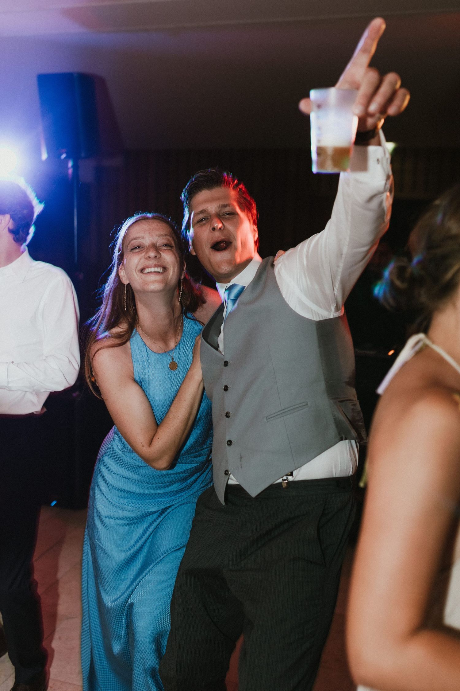 Guests dancing and celebrating at lively wedding reception with colorful lighting.