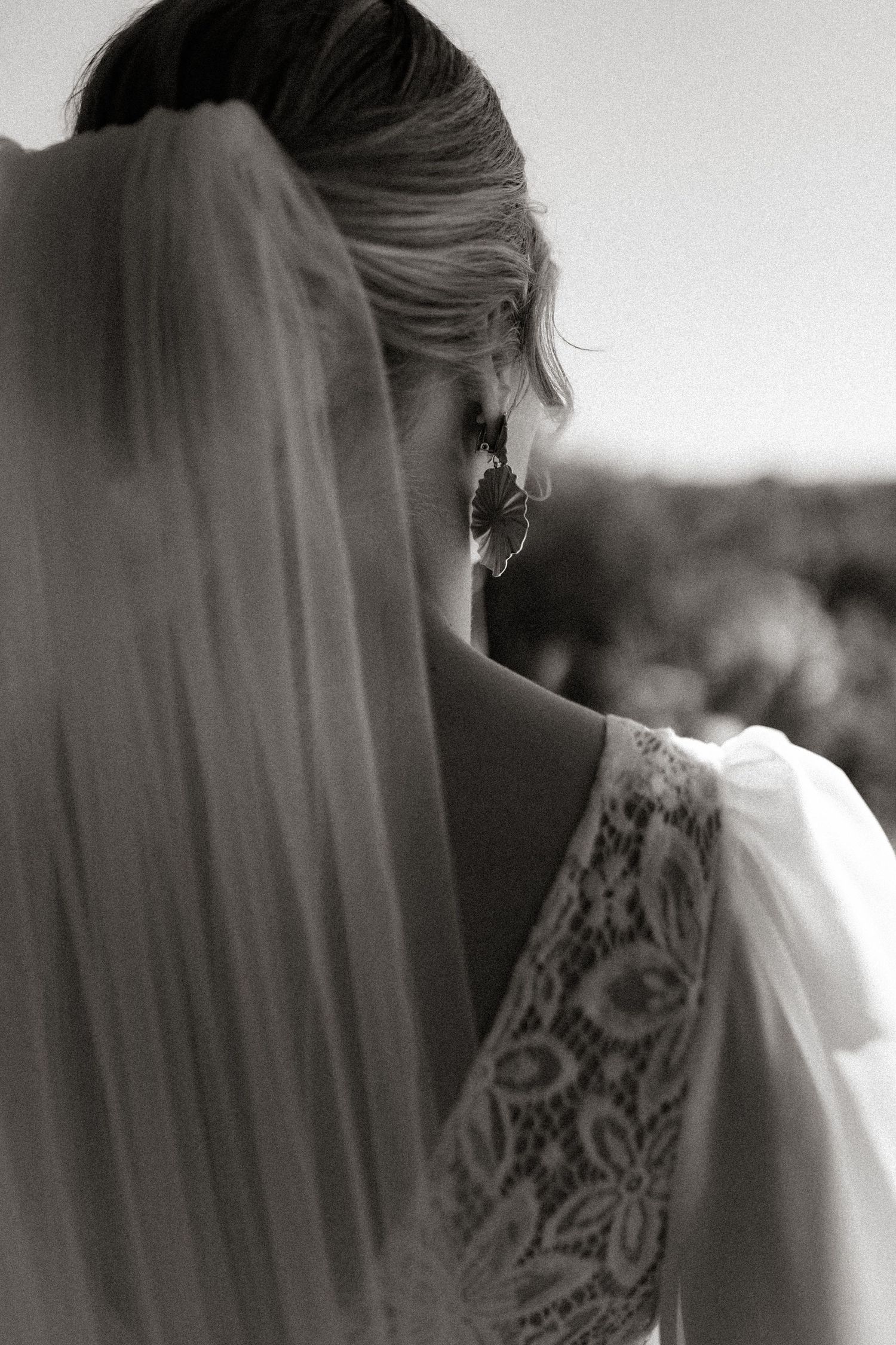 Black and white detail shot of a bride's lace wedding dress and flowing veil against a blurred background.
