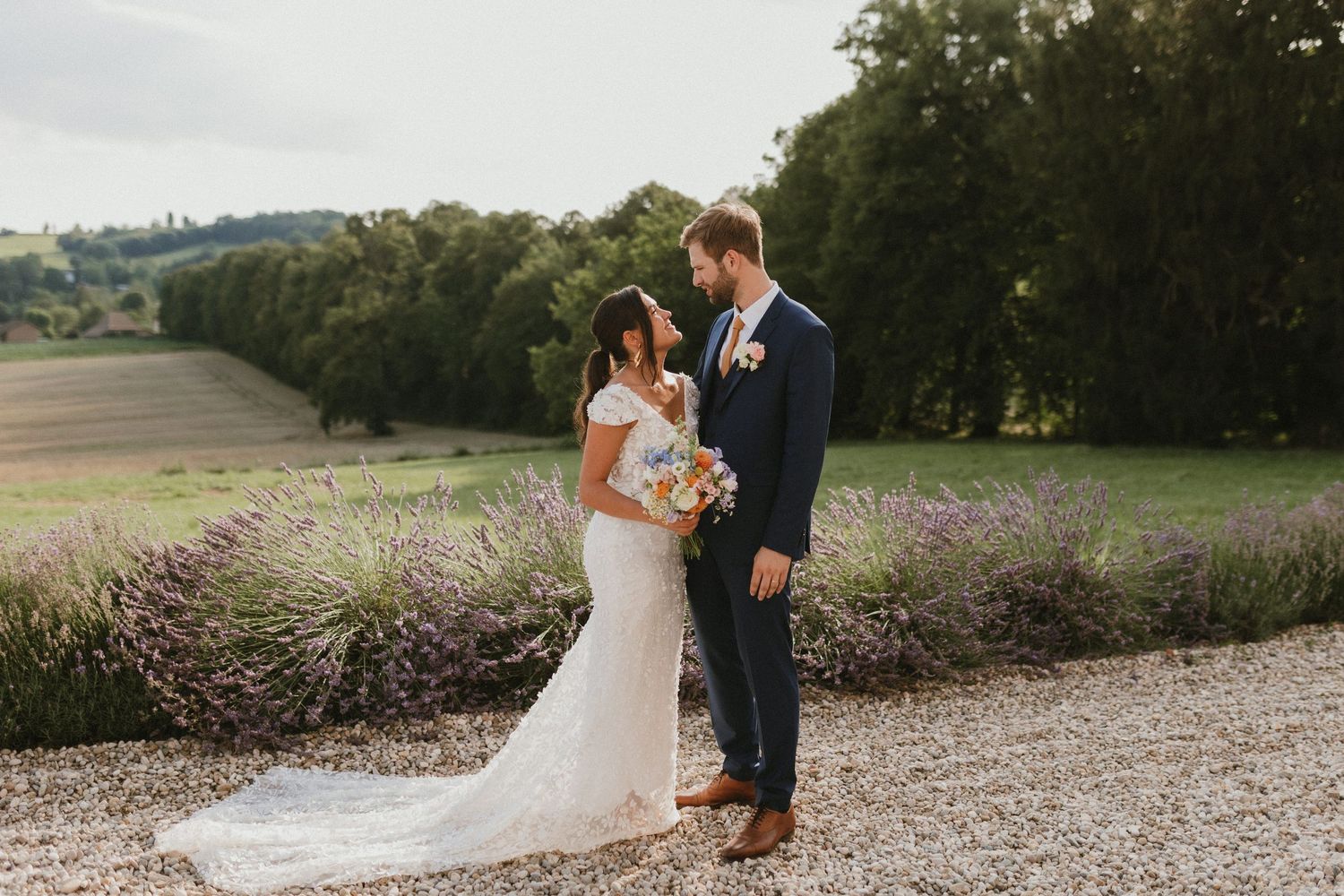 A couple stands together on a gravel path with scenic countryside views and lavender fields.