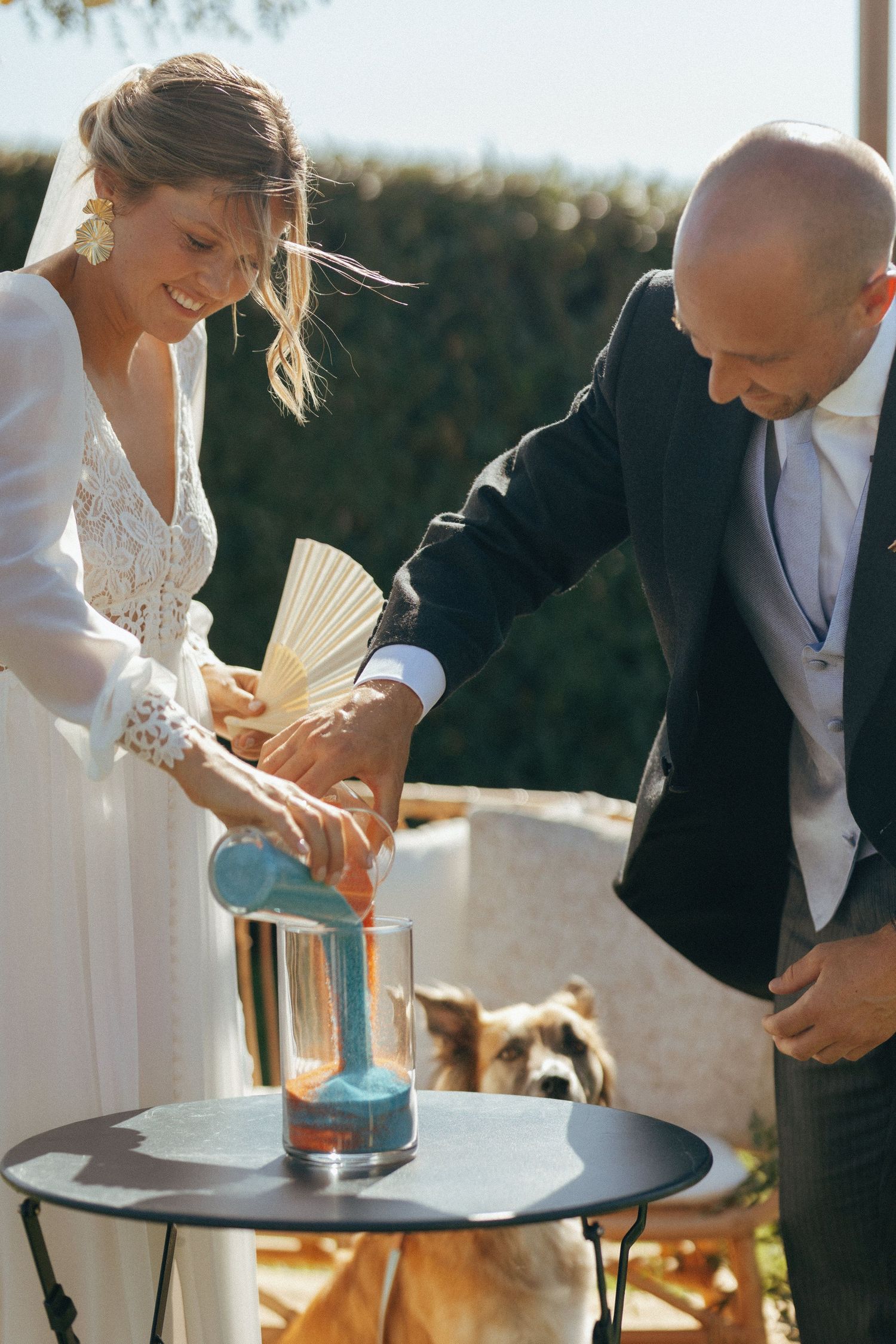 Couple performs unity ceremony at outdoor wedding by pouring colored sand into glass vessel.