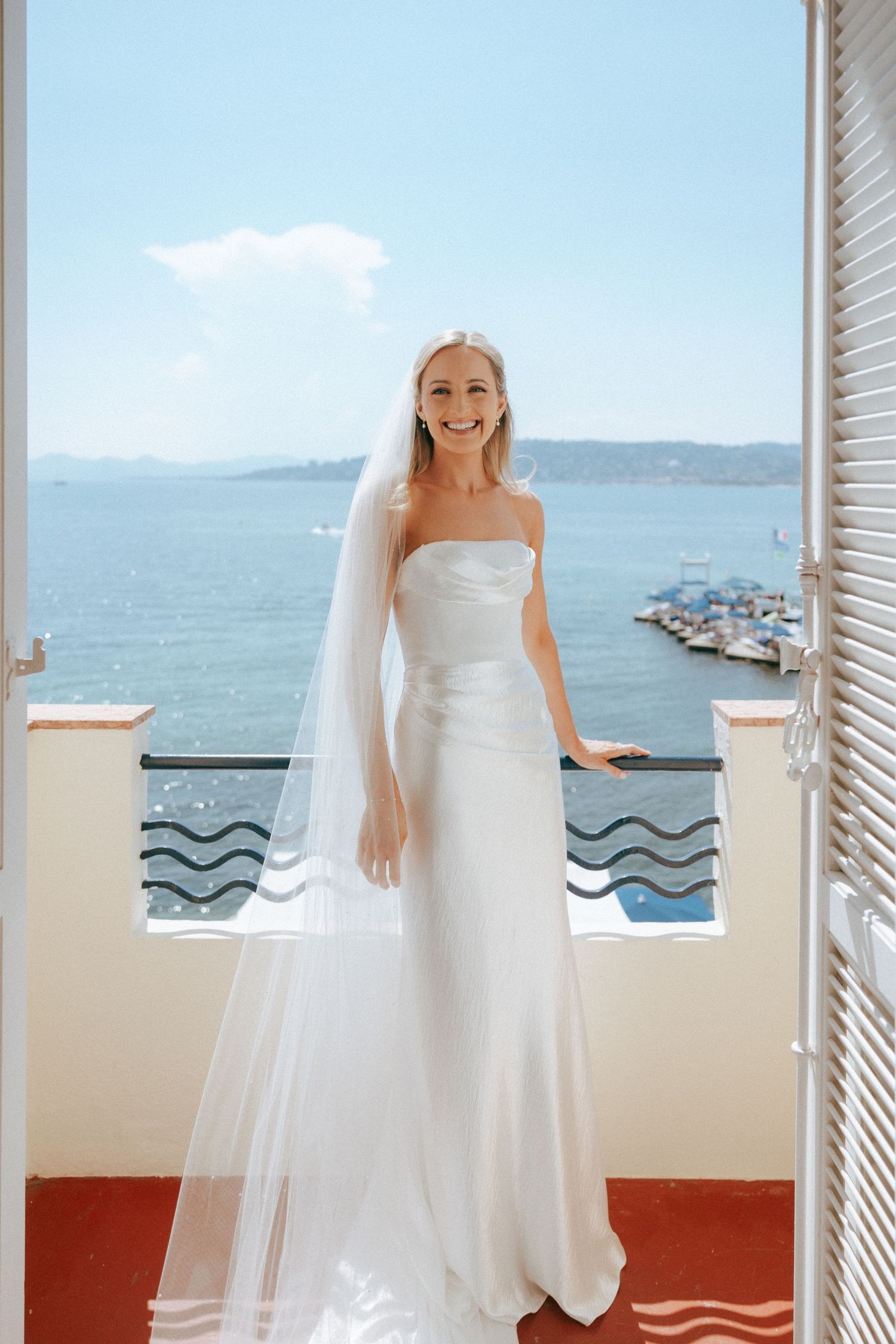 Elegant bride in a classic white strapless wedding gown stands on a balcony overlooking a scenic marina and ocean view.