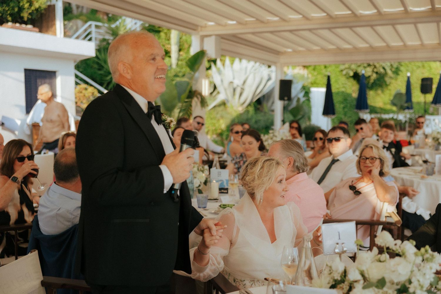 Elegant white floral centerpieces decorate tables at an outdoor wedding reception under a pergola.