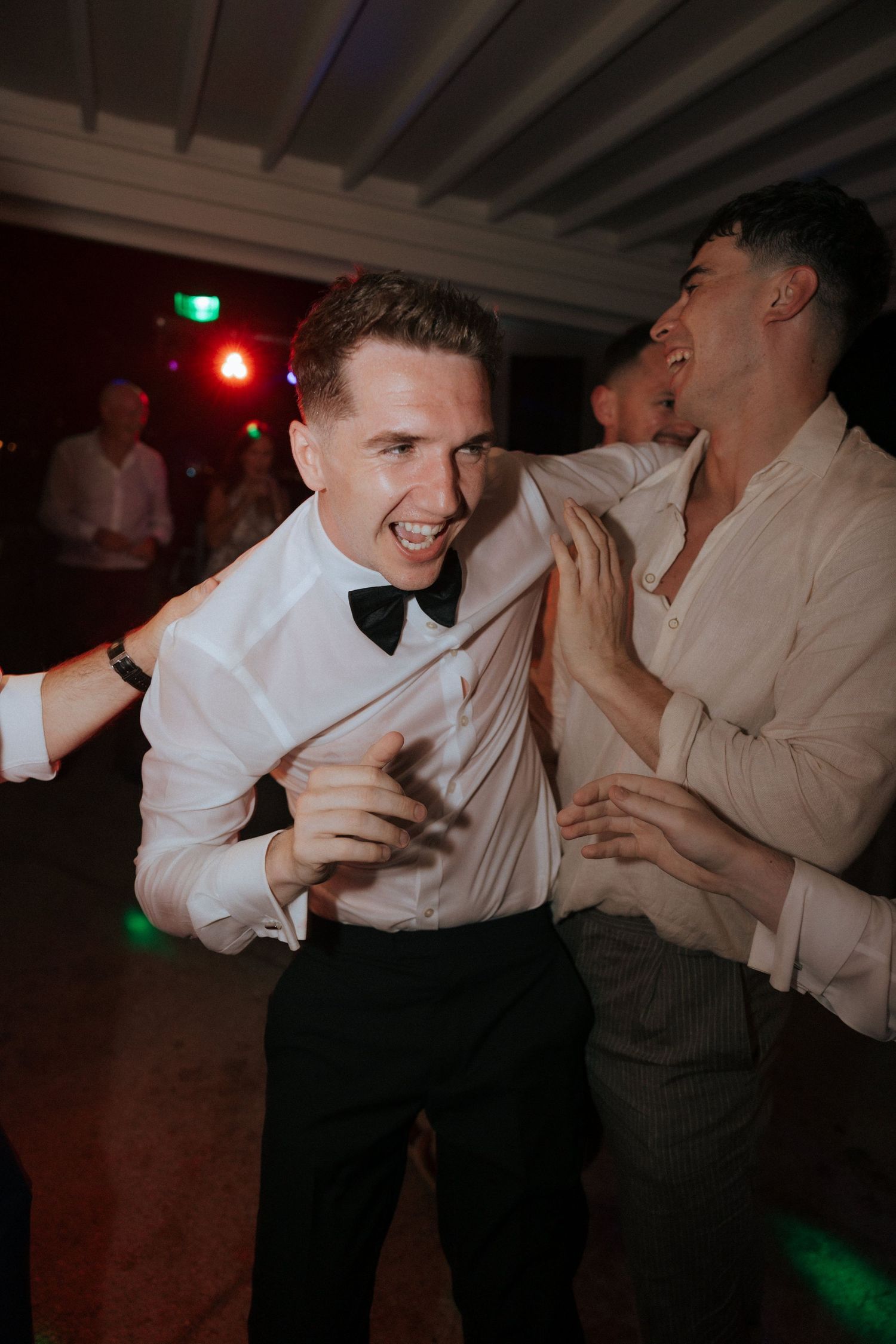 A guest in formal attire dances enthusiastically at a wedding reception under colored lighting.