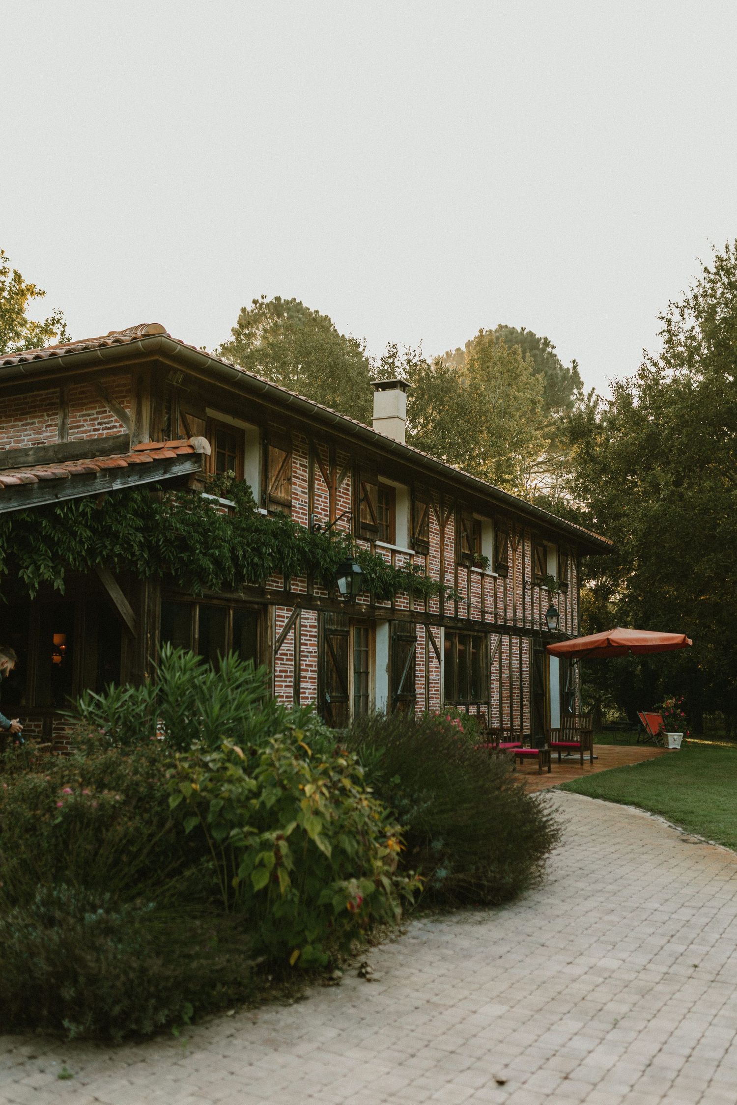 Rustic brick ranch house with large windows and lush landscaping along a stone paved walkway at dusk.