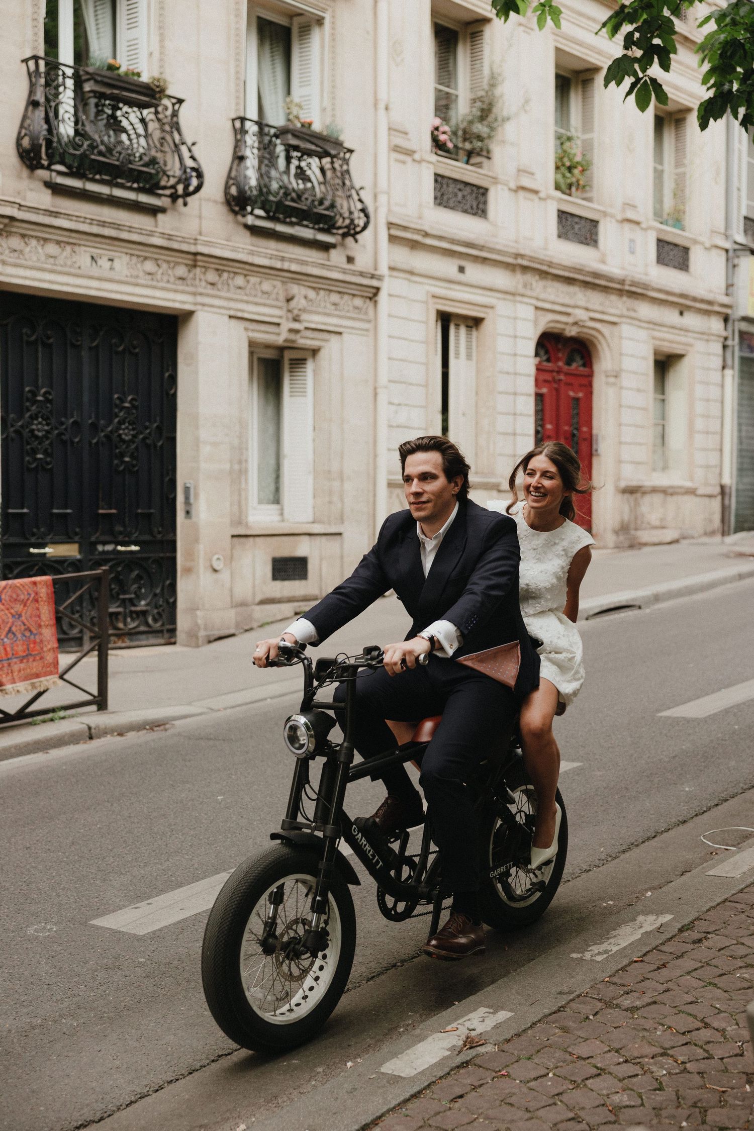 A couple rides a vintage motorcycle down a charming Parisian street with classic architecture and ornate balconies.