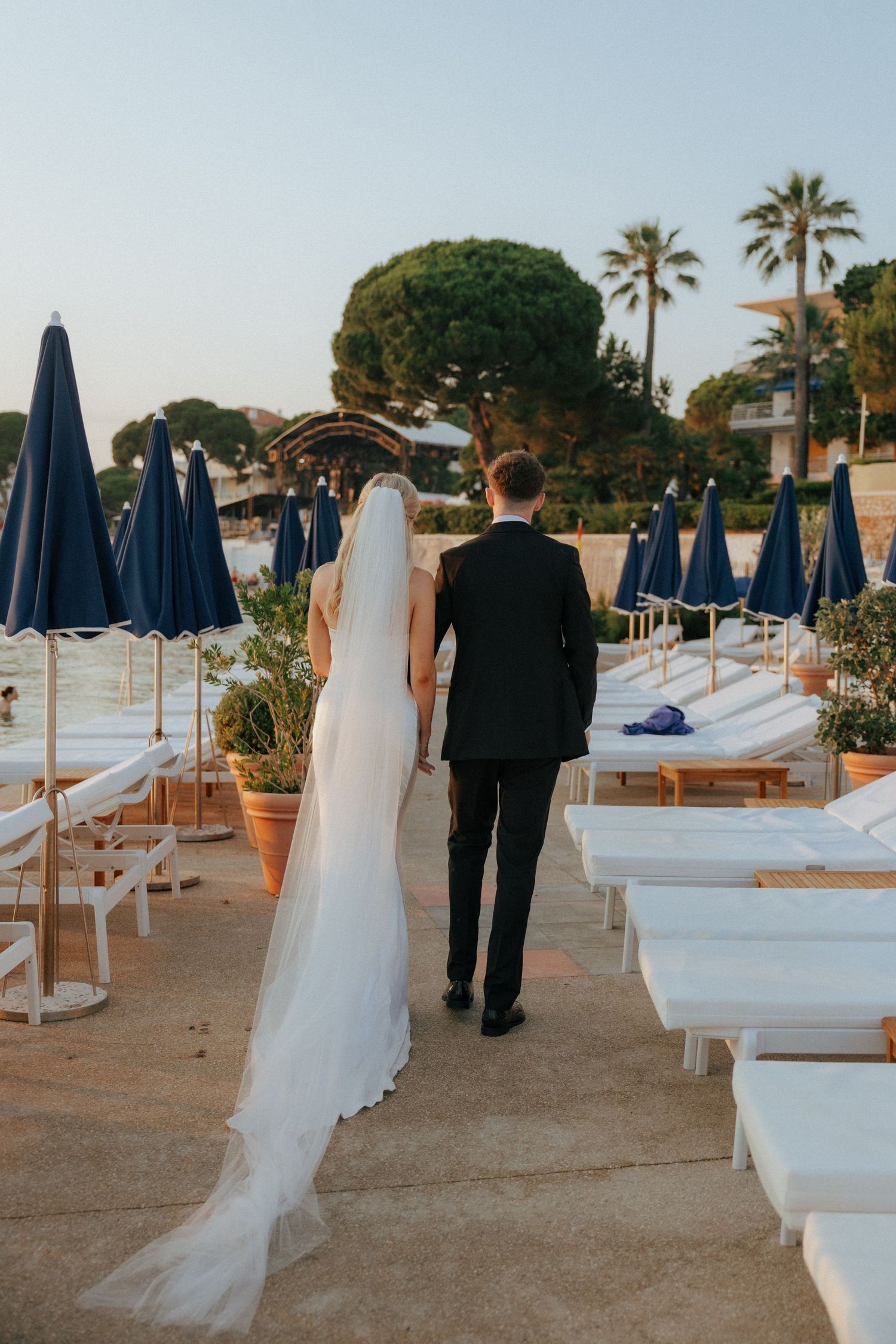 Wedding couple walking through beach club setting with white lounge chairs and blue umbrellas.