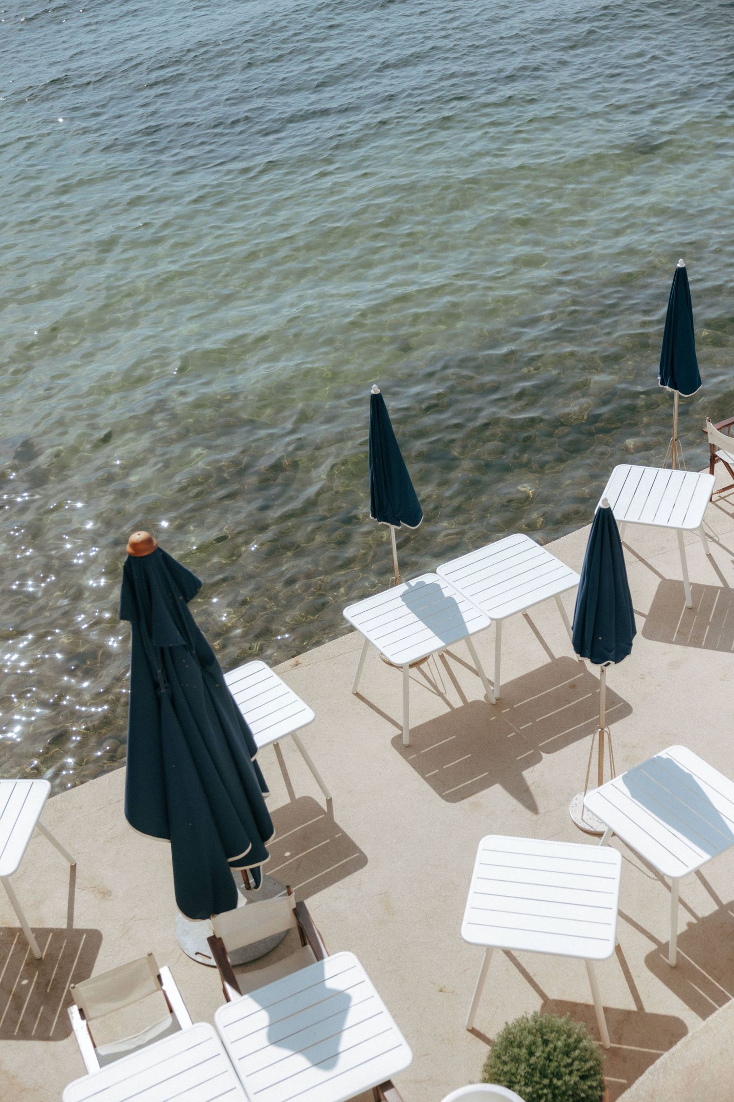 Aerial view of beach deck with white lounge chairs and navy umbrellas overlooking sparkling water.