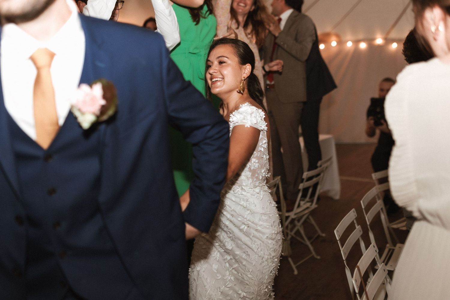People dancing and celebrating at a wedding reception under a tented venue with string lights.