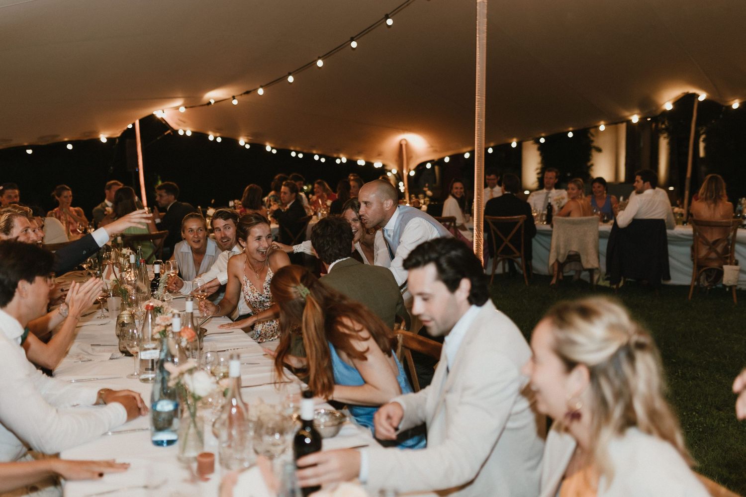 Wedding guests gather under string lights at long tables during an outdoor evening reception.