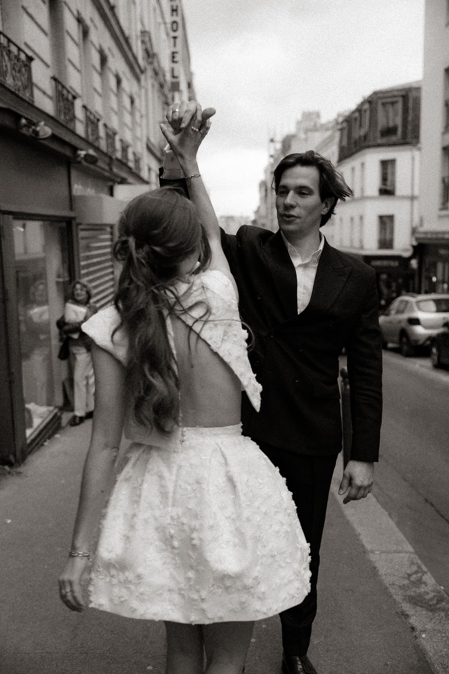 Black and white street photography captures a couple in wedding attire walking along an urban street in Paris.