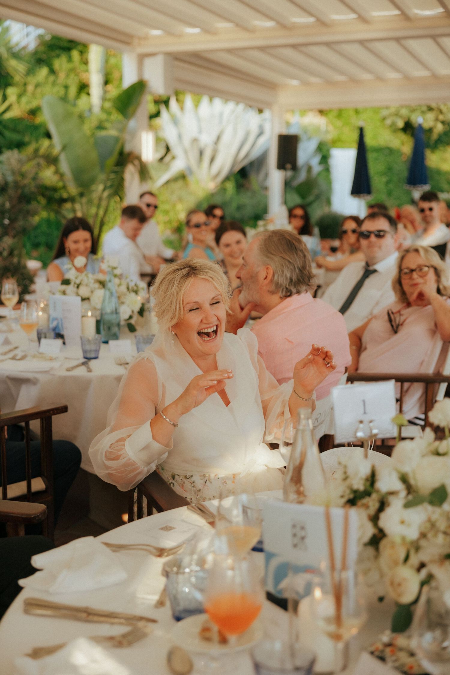 Wedding guests enjoy a lively reception meal under an outdoor pergola with floral centerpieces.