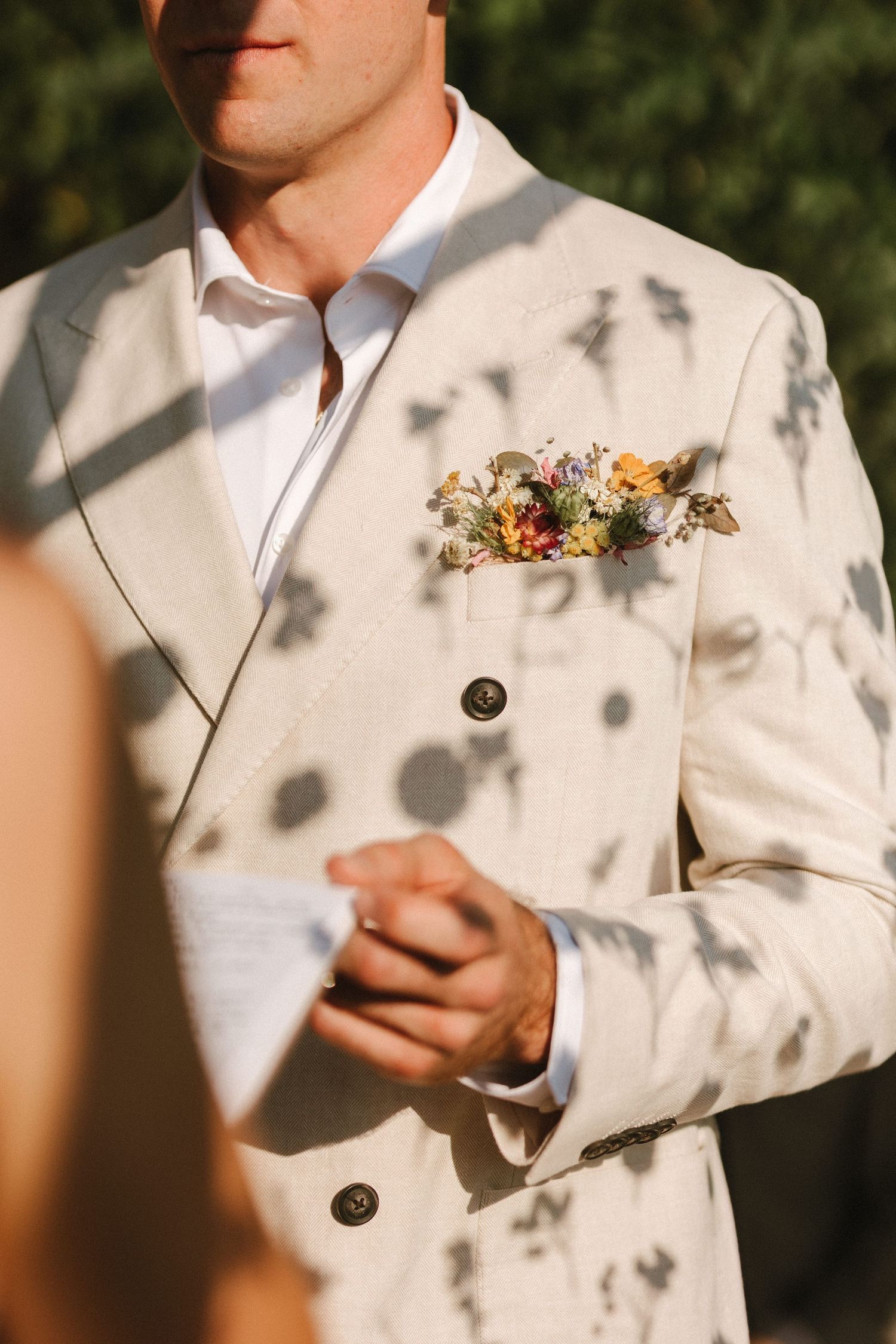 A floral boutonniere adorns a cream-colored suit jacket casting dappled shadows in sunlight.