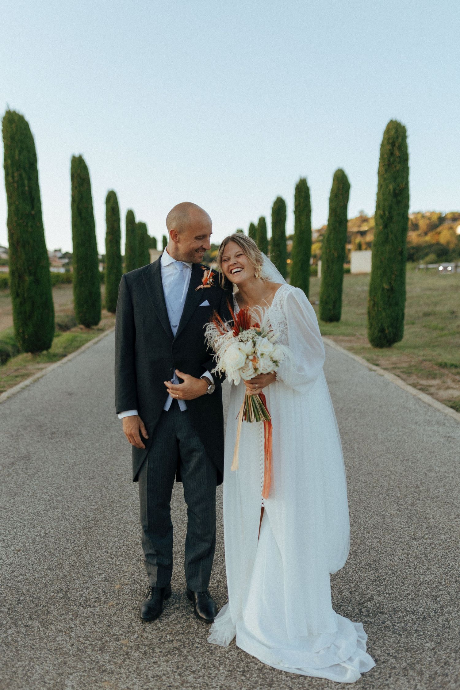 Newlyweds share joyful moment on cypress-lined road in elegant wedding attire.