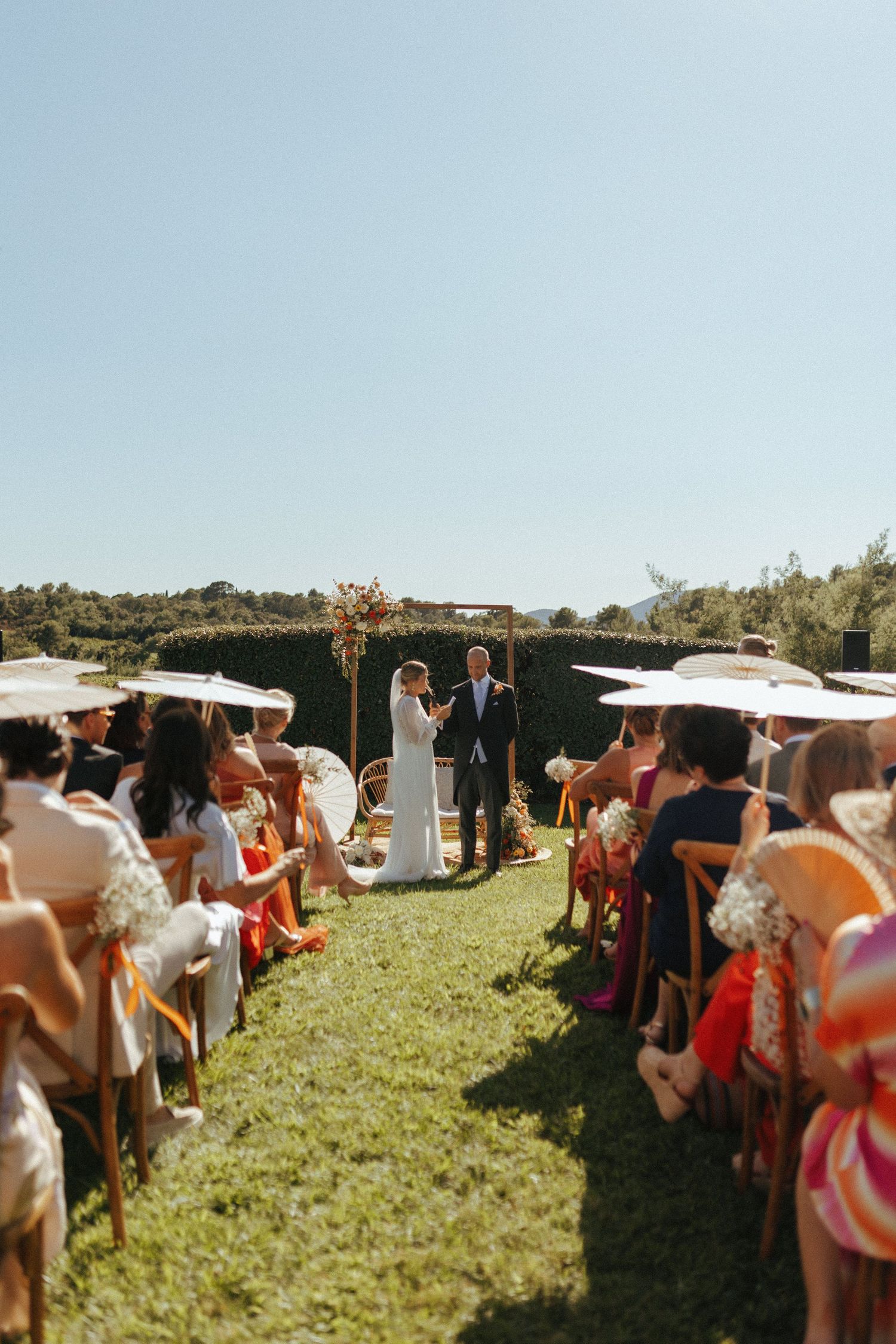 An outdoor wedding ceremony takes place on a grassy hill under blue skies with guests seated on white chairs.
