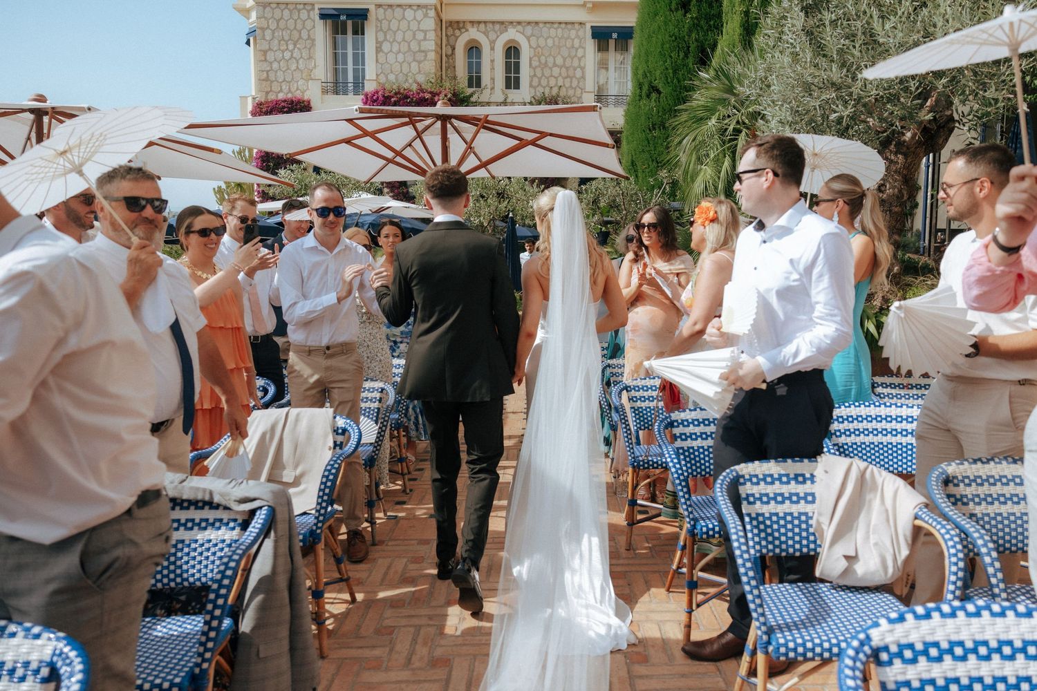 Guests gather at a Mediterranean-style outdoor restaurant with blue checkered chairs and white umbrellas on a sunny day.