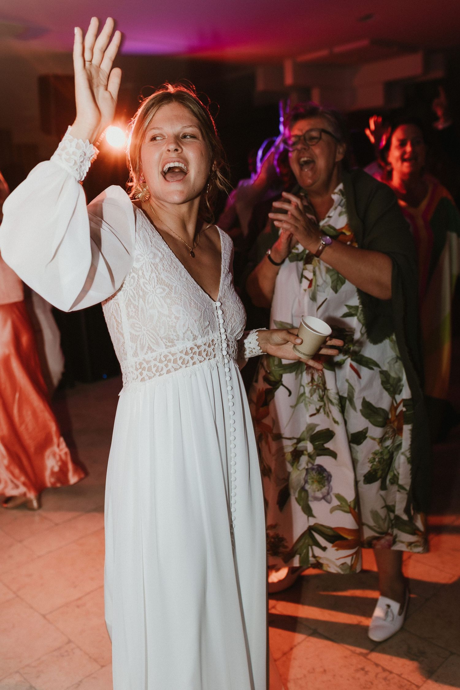 Bride in white dress celebrating on dance floor with guests at wedding reception in colorful lighting.