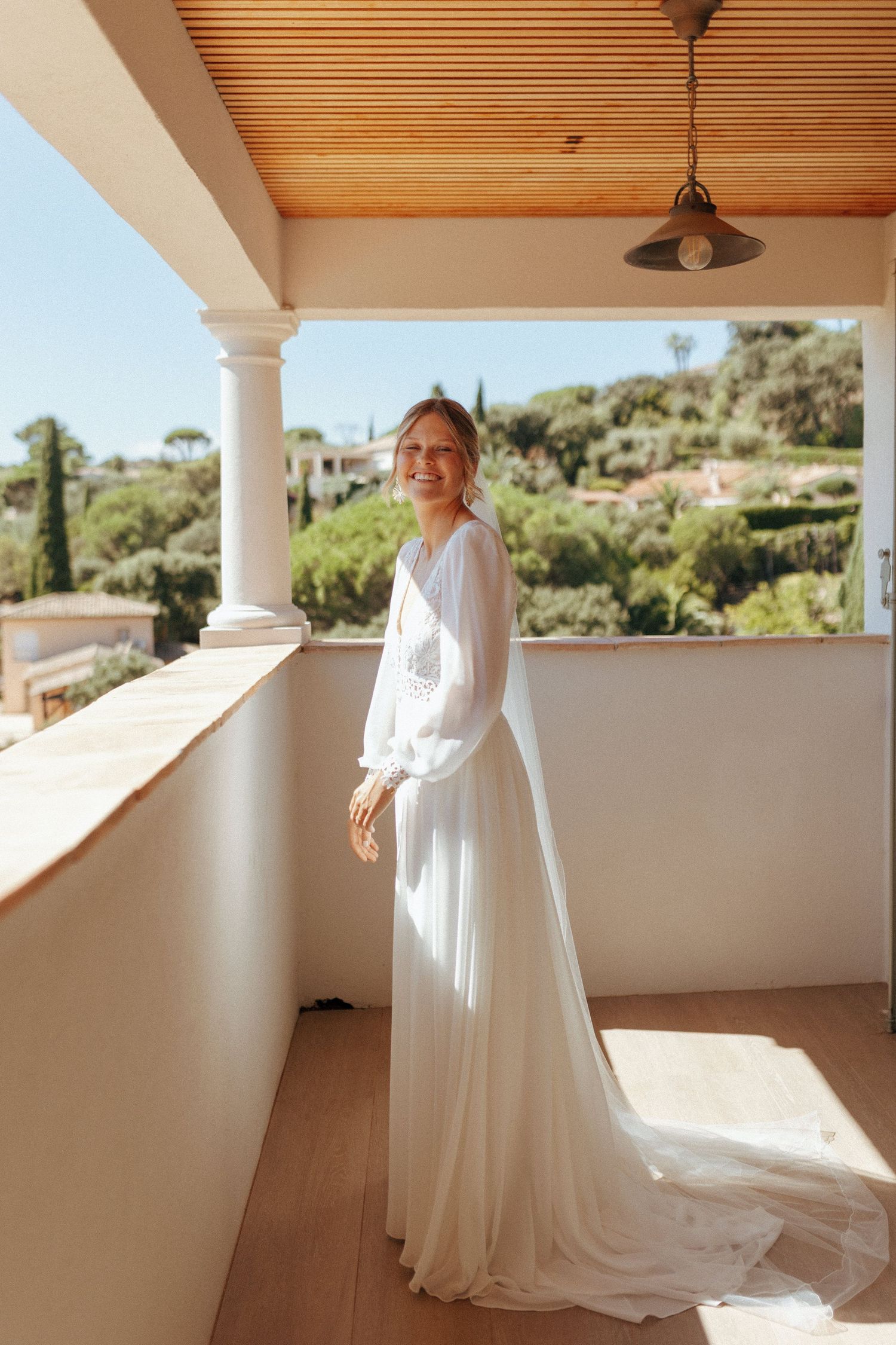 Elegant white wedding dress with long sleeves photographed on a Mediterranean-style balcony with wooden ceiling and column.