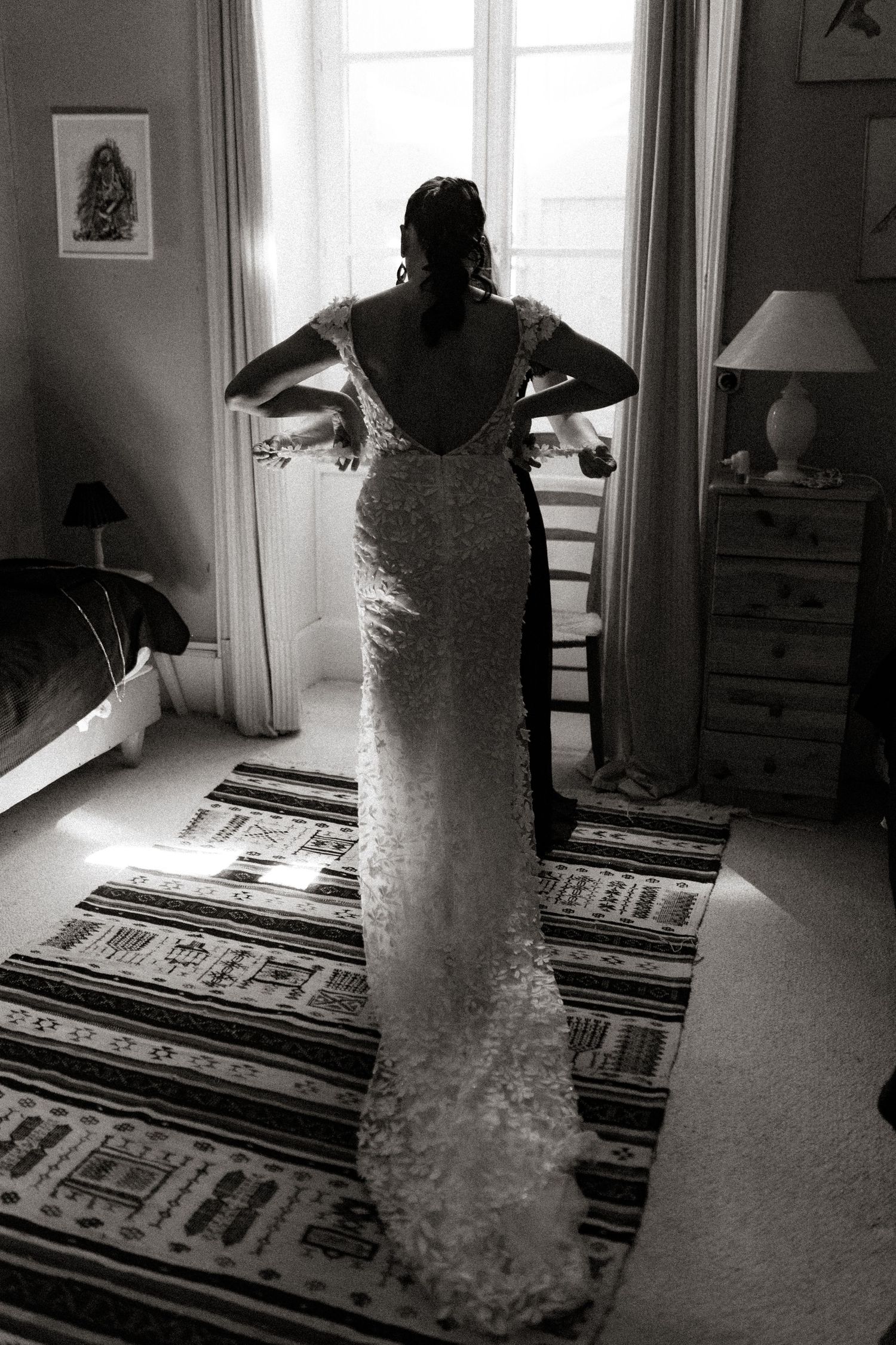 A bride in a backlit silhouette stands by a window wearing a fitted lace wedding gown in a black and white photograph.