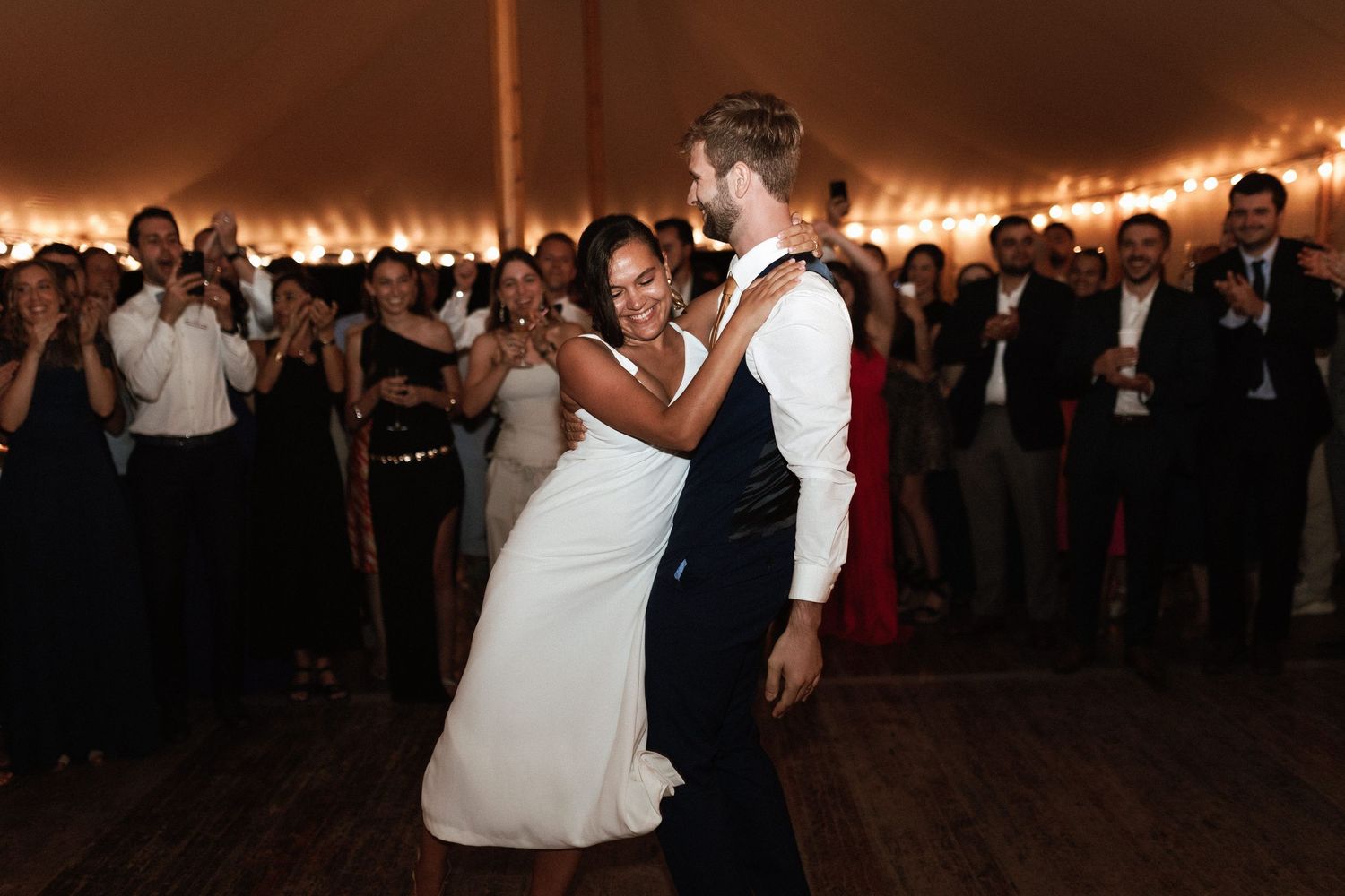 Wedding couple shares an intimate dance moment surrounded by guests under warm tent lighting.