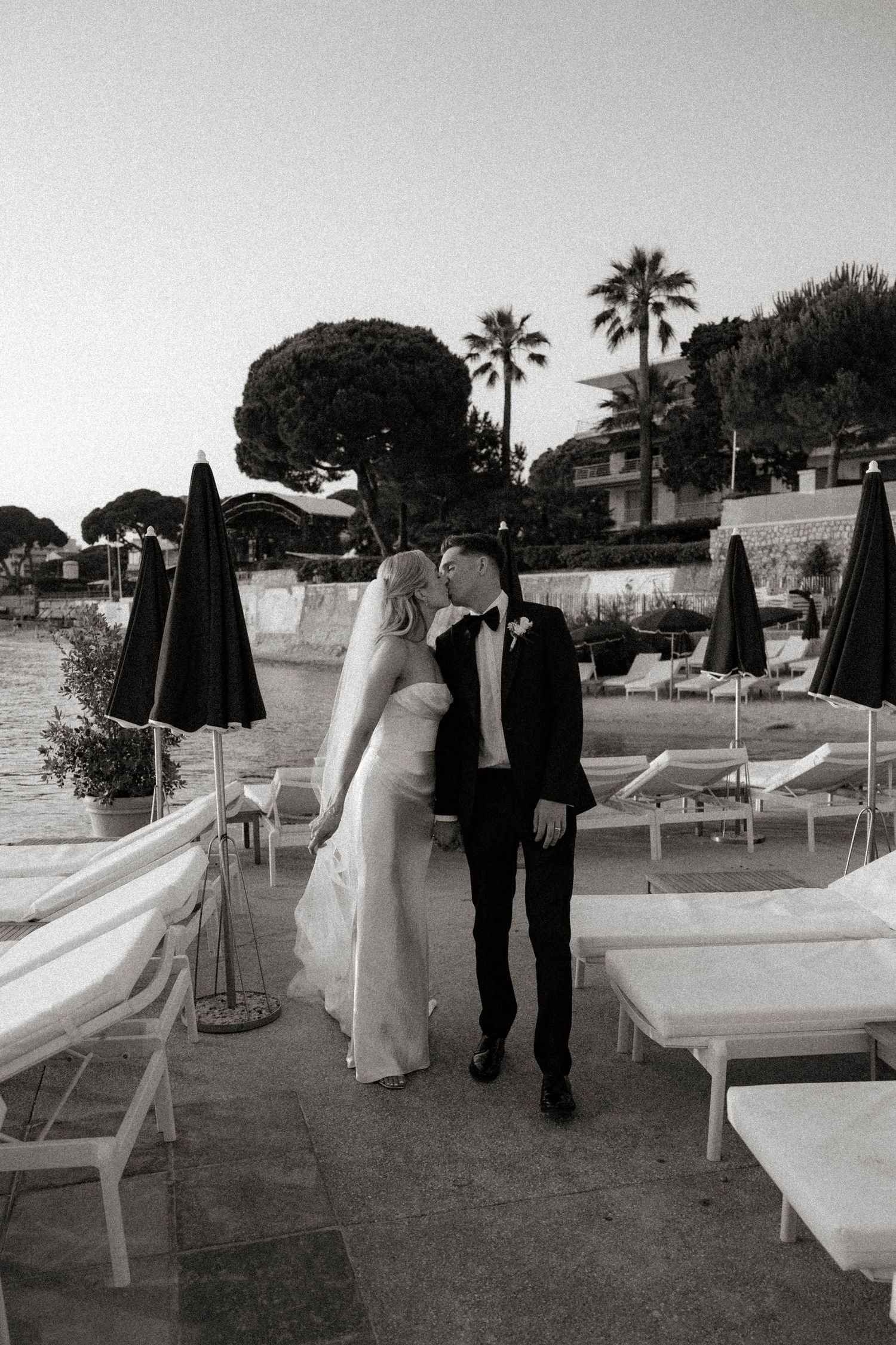 A romantic black and white wedding photo of a couple embracing on a resort patio with lounge chairs and palm trees.