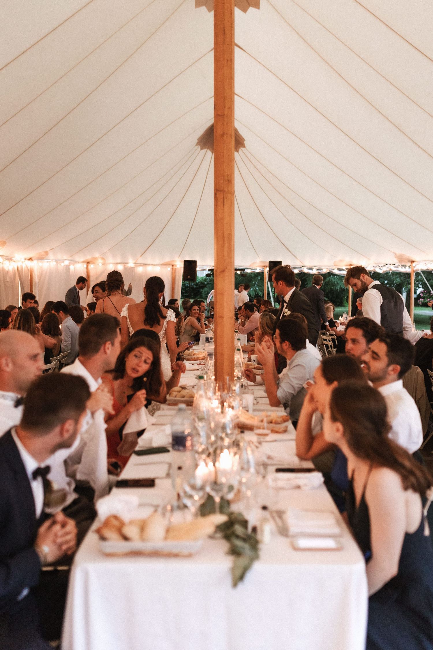 Wedding guests seated at elegant long tables under a white reception tent with candlelight and draped fabric decor.