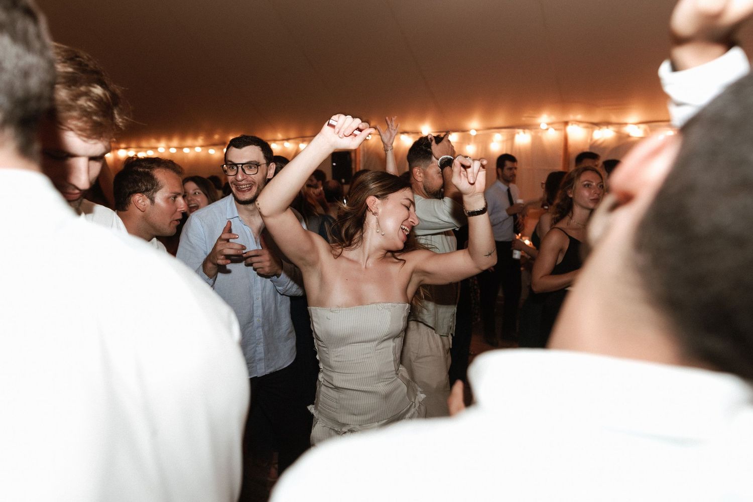 Celebration on a dance floor with guests dancing under string lights at wedding reception.