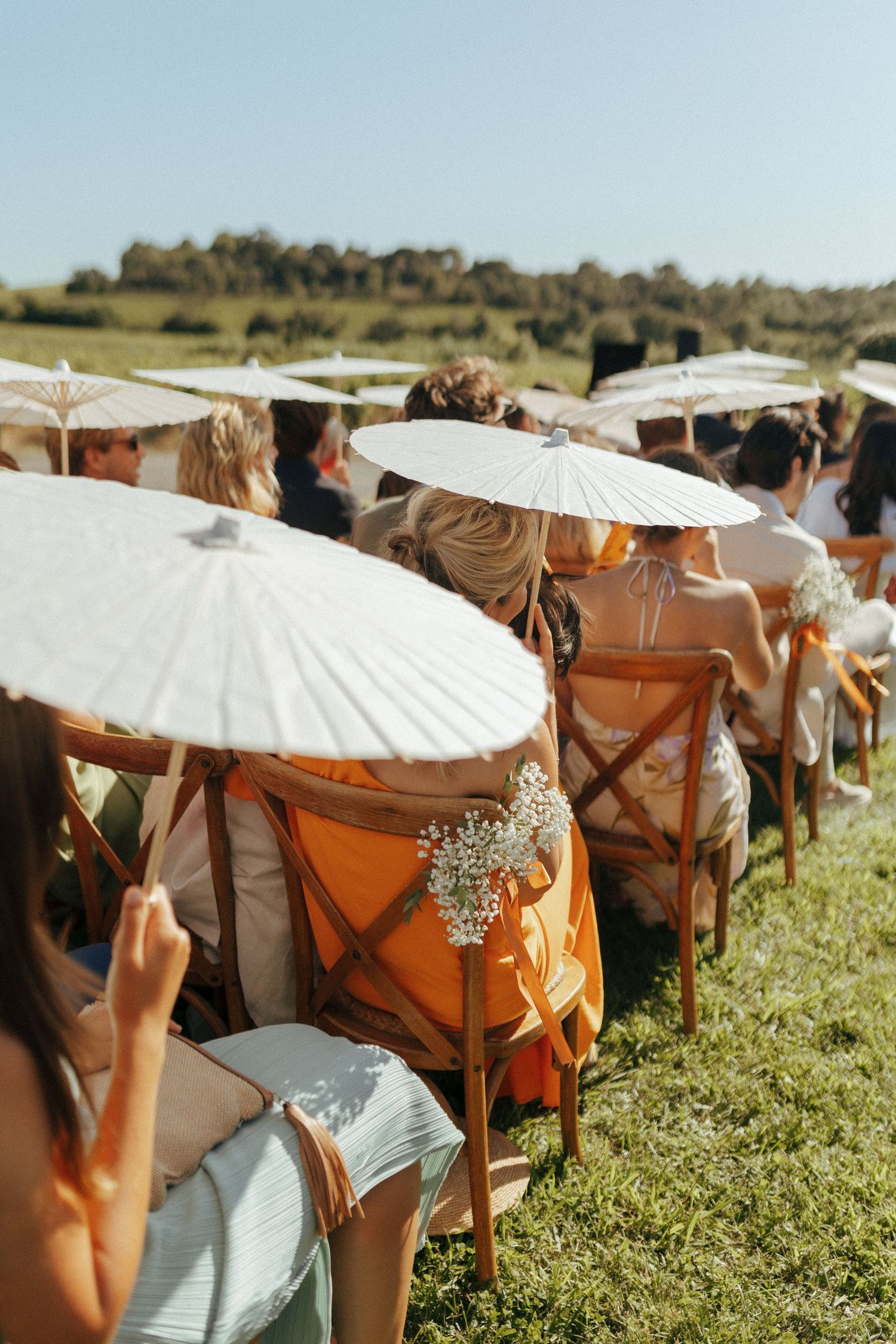 Wedding guests seated in wooden chairs holding white parasols during an outdoor ceremony.