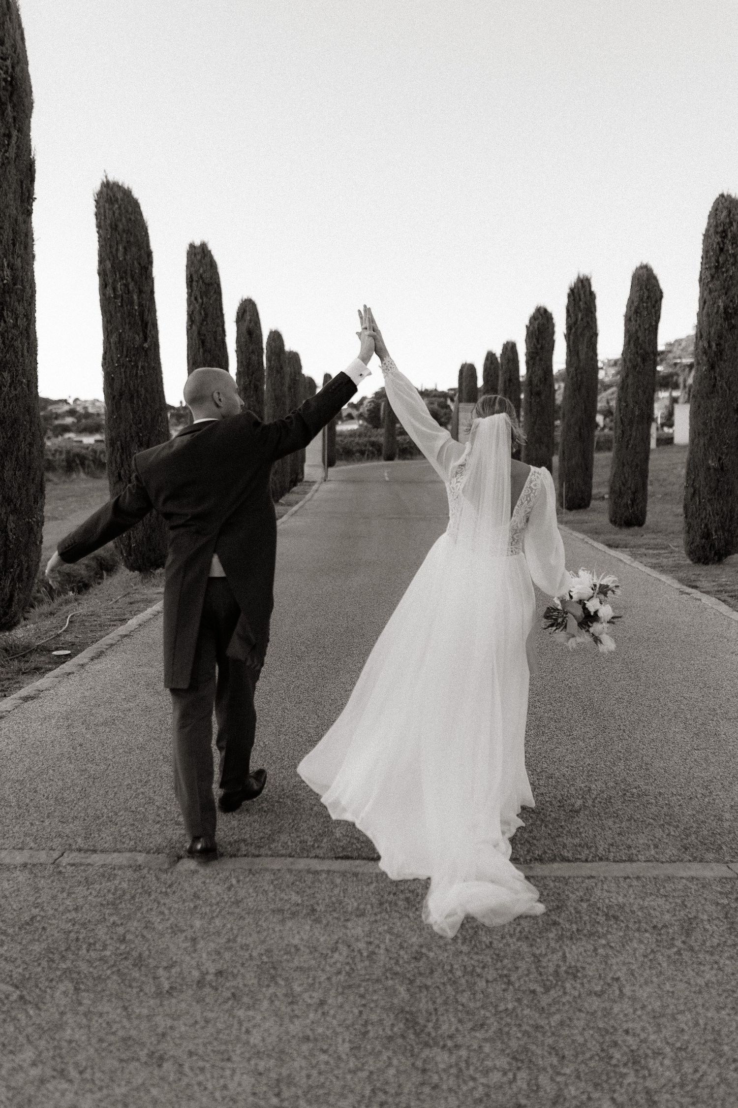 Black and white photo of newlyweds celebrating with high fives along cypress-lined path.