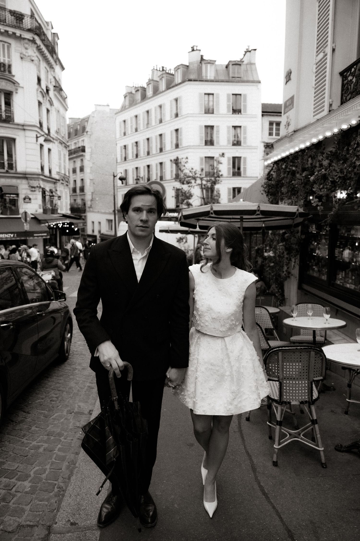 A stylish couple walks along a charming Parisian street with classic architecture and sidewalk cafes in the background.