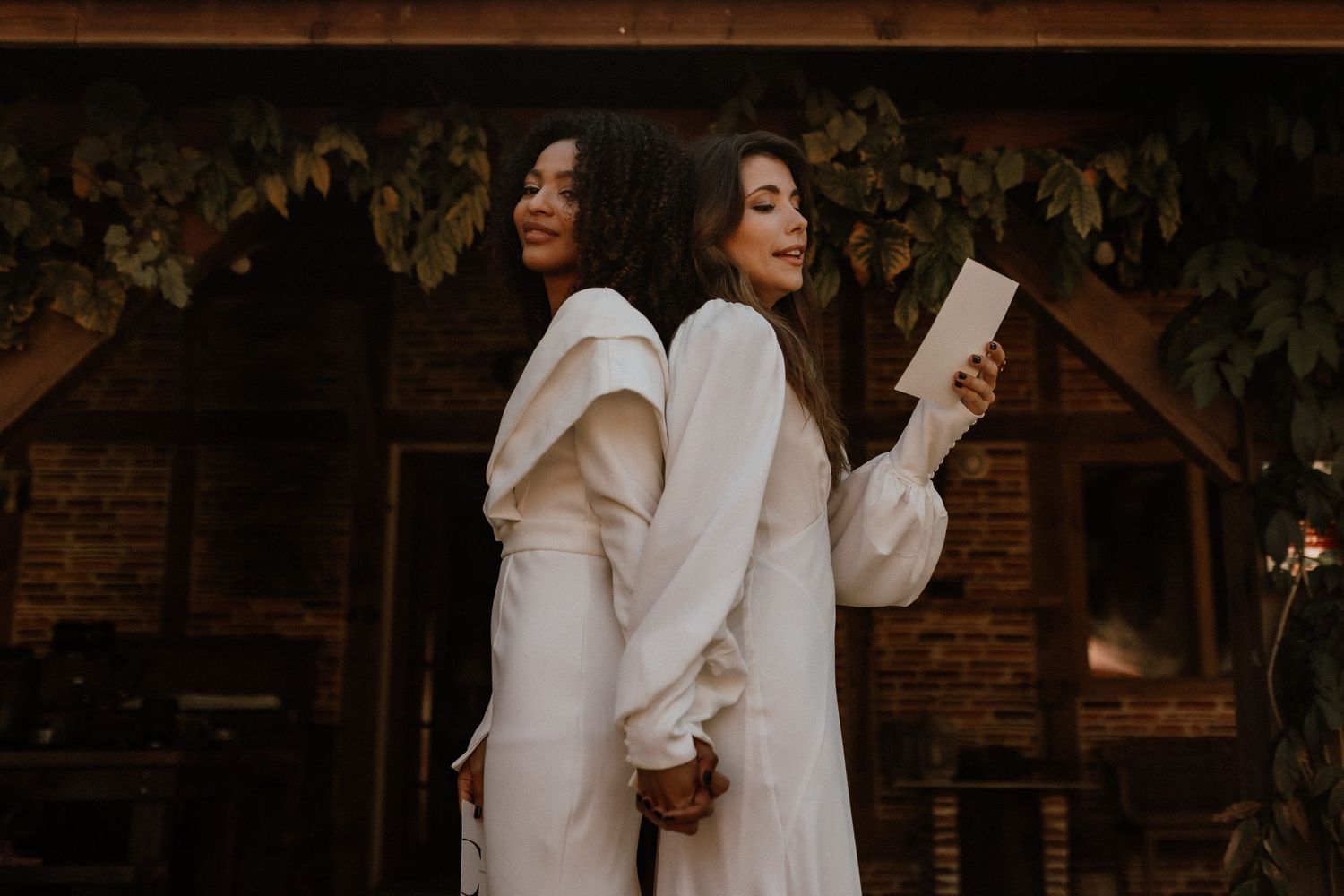 Two people wear elegant white dresses while standing back to back in front of a rustic wooden structure.